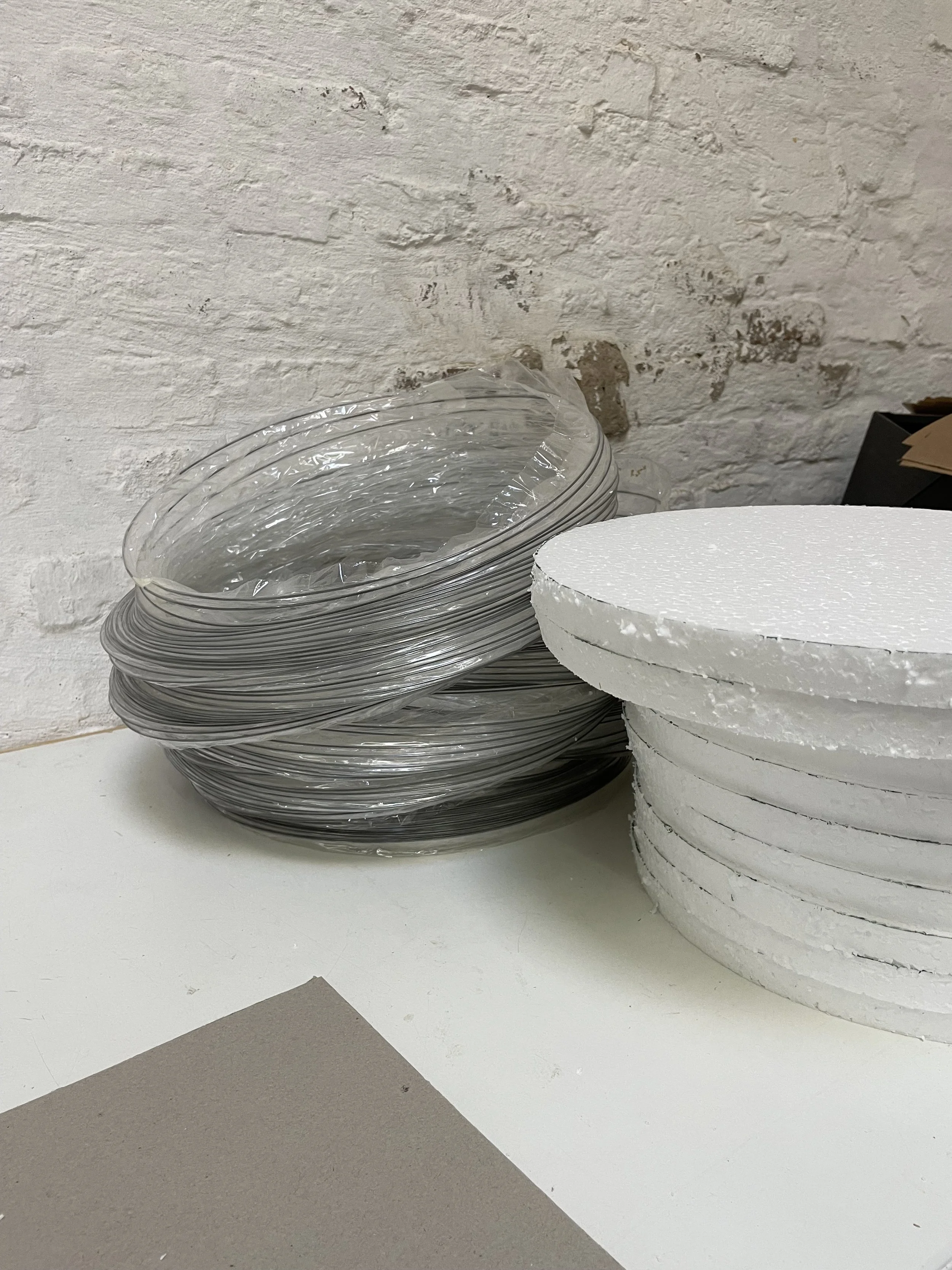 Stack of clear plastic plates wrapped in plastic next to a stack of white disposable plates on a white table against a white brick wall.