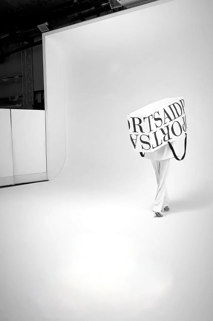 Person holding a large shopping bag with partially visible text, standing in a studio with a plain white background.