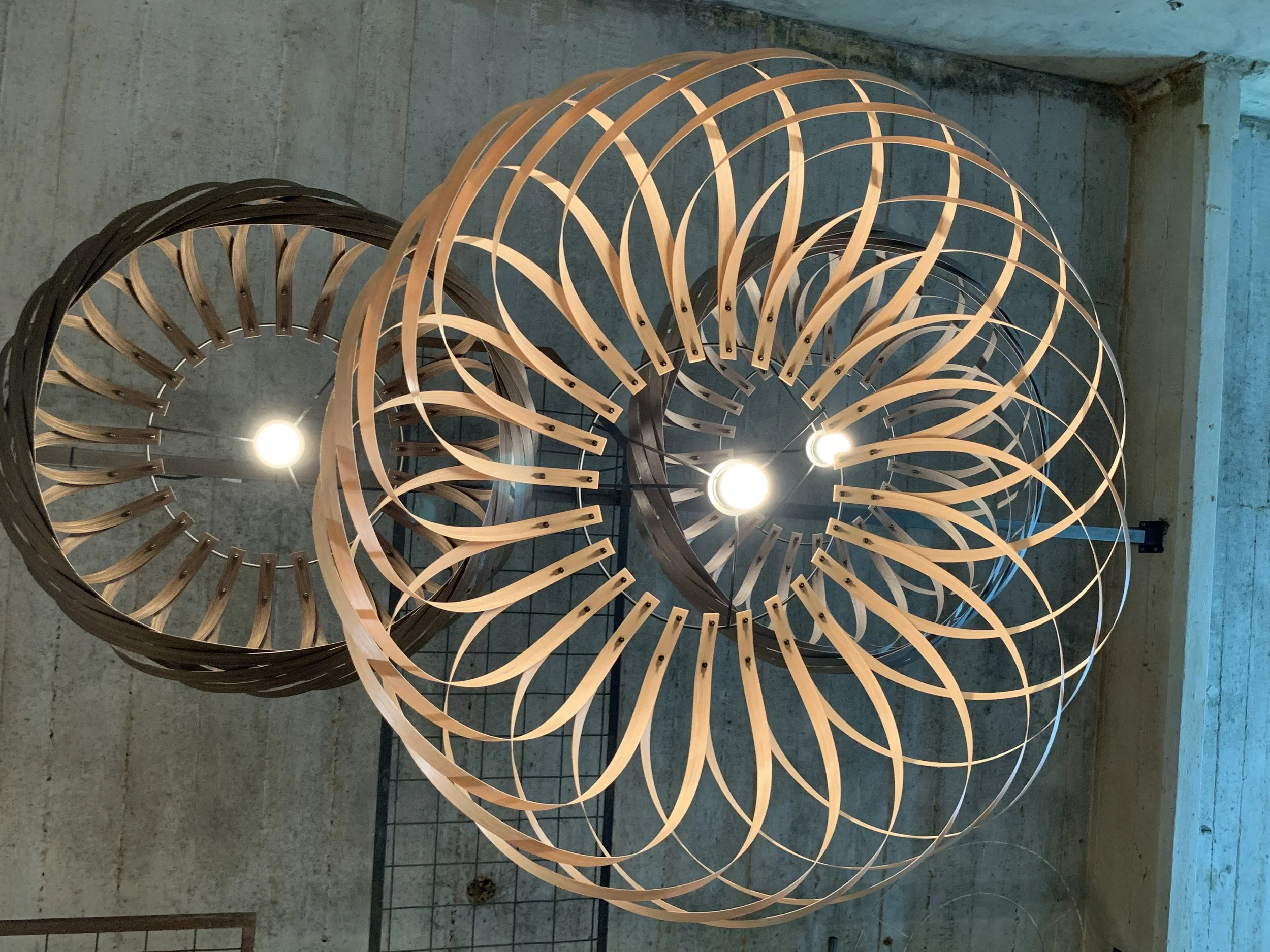 Three modern chandelier light fixtures made of wooden frames with intricate geometric patterns, hung at different heights from the ceiling of an industrial-style concrete space.