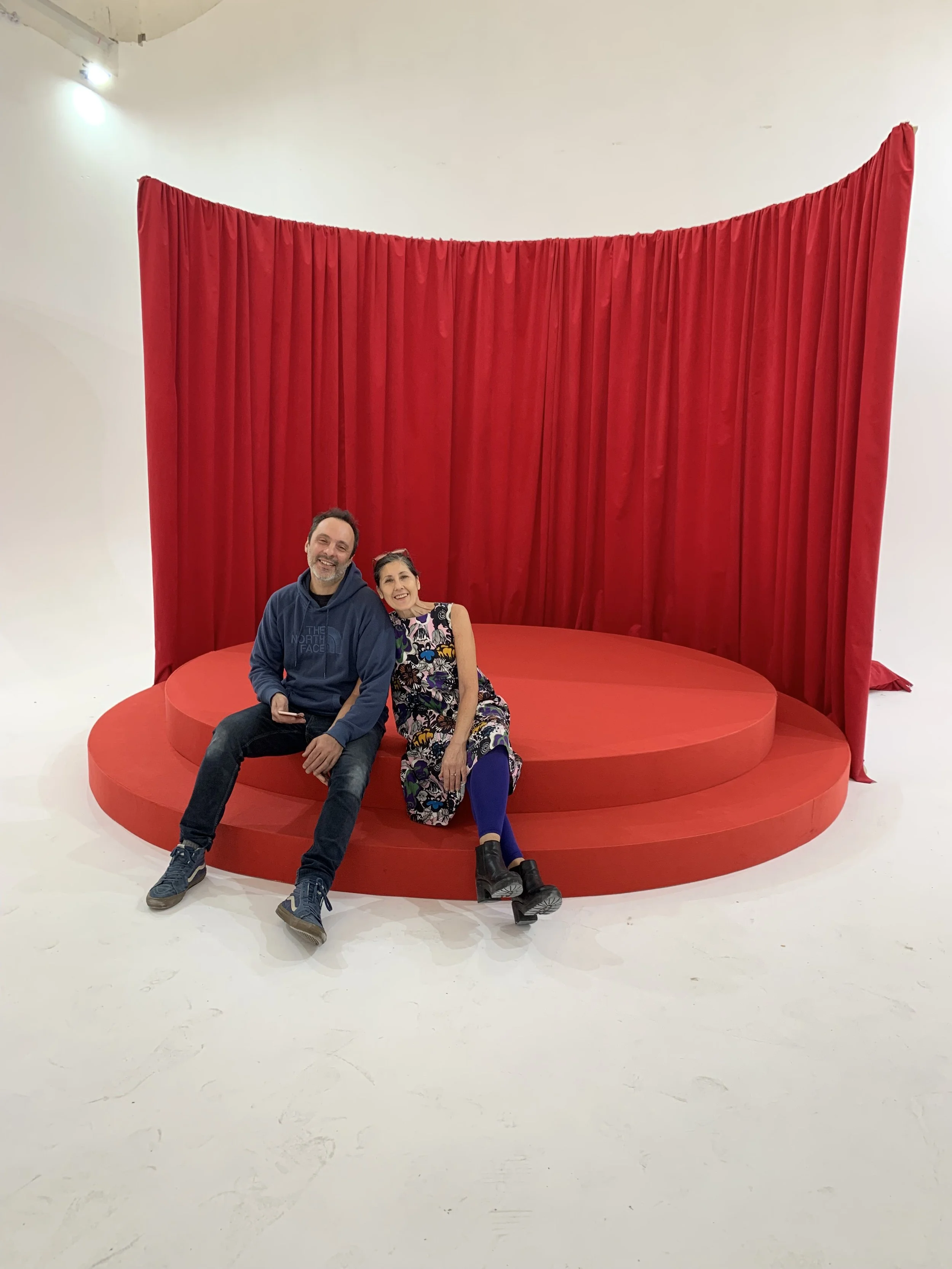 Two people sitting on a round red stage with a tall red curtain backdrop, smiling and posing for the photo.