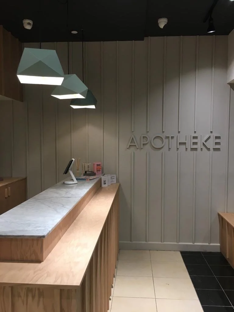 Interior of a modern pharmacy with a wooden counter and a white marble top, a digital cash register, and a wall with the word 'APOTHEKE' in large letters.