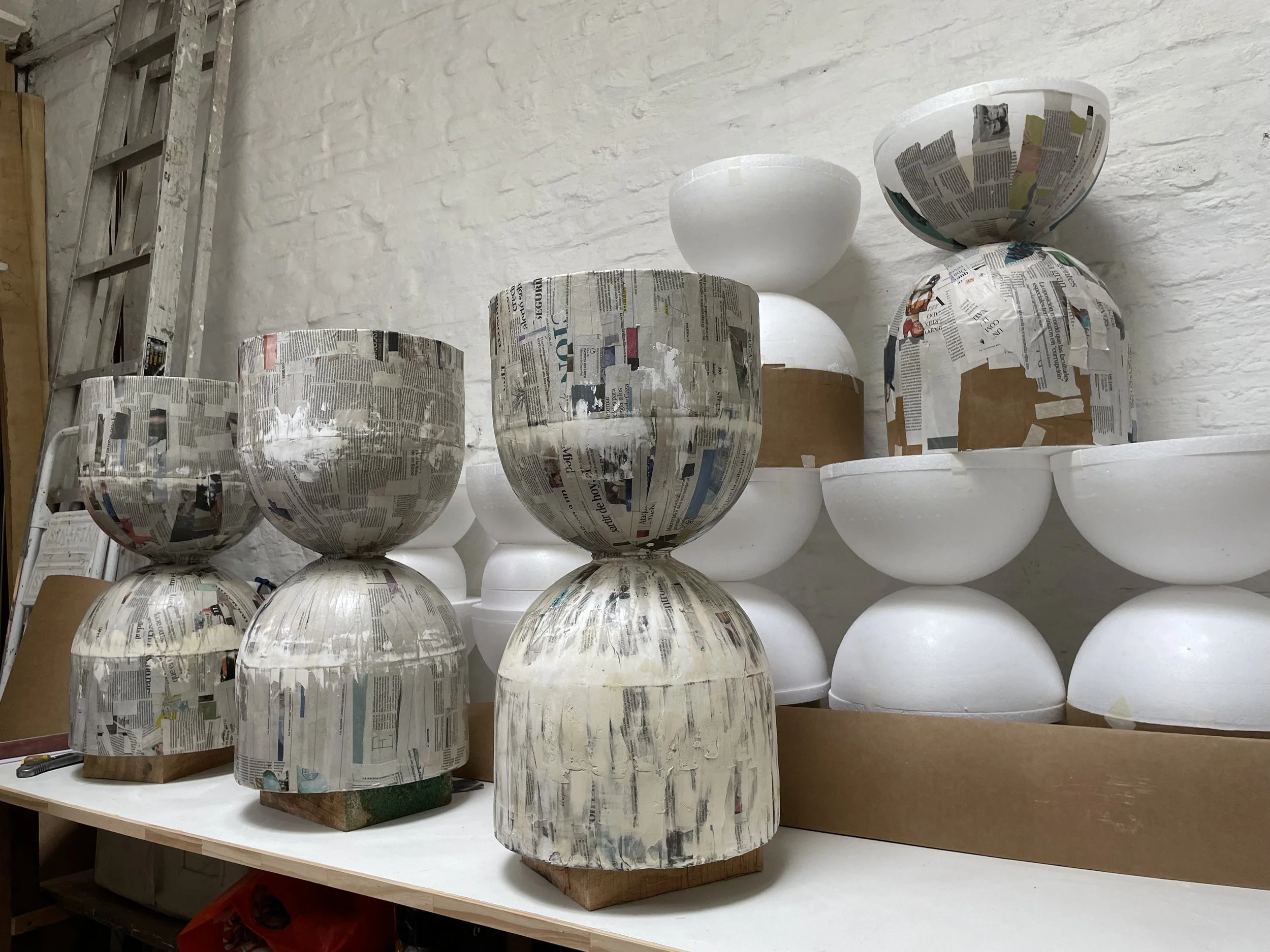 Several large bowls and vases in various stages of being painted or decorated, covered with newspaper, on a worktable against a white brick wall.