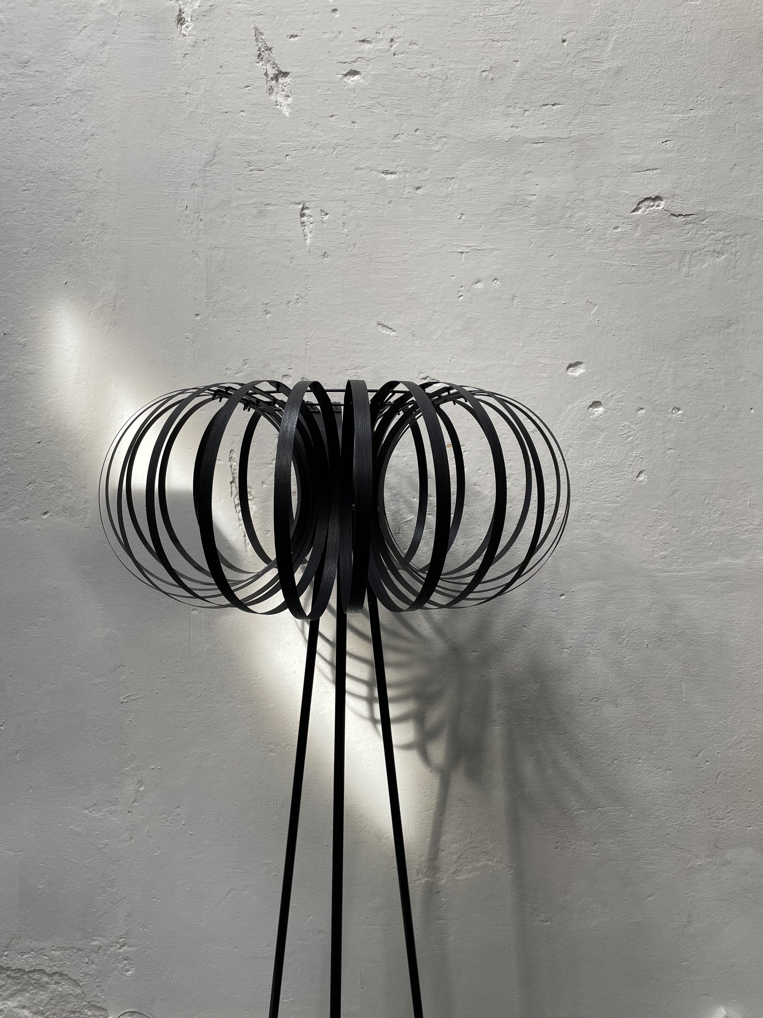 Black metal sculpture with circular and vertical elements casting shadows on a textured white wall.