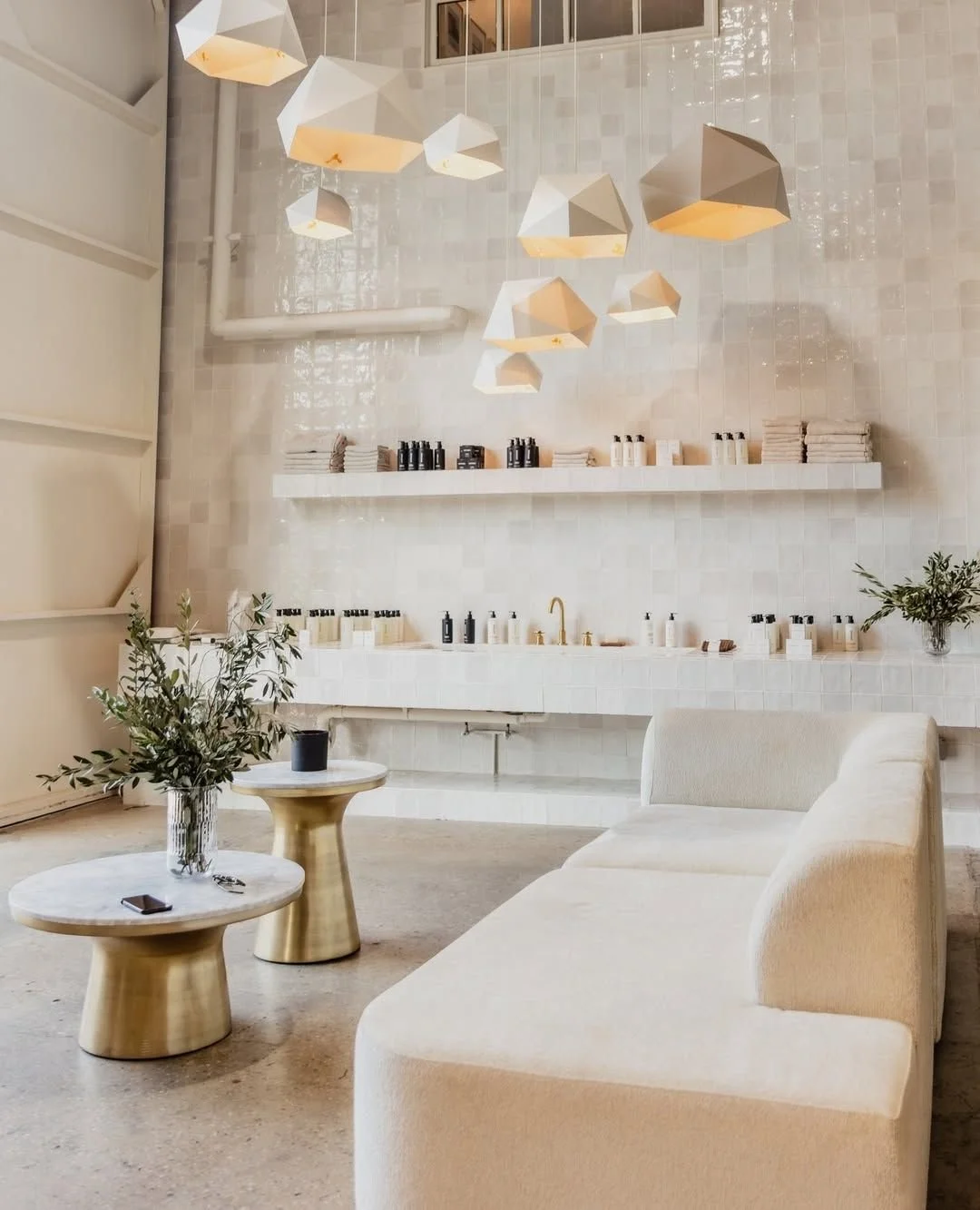 Modern minimalist interior with white tiled walls, hanging geometric pendant lights, a white sofa, marble-top tables with gold bases, and a shelf with skincare or beauty products.