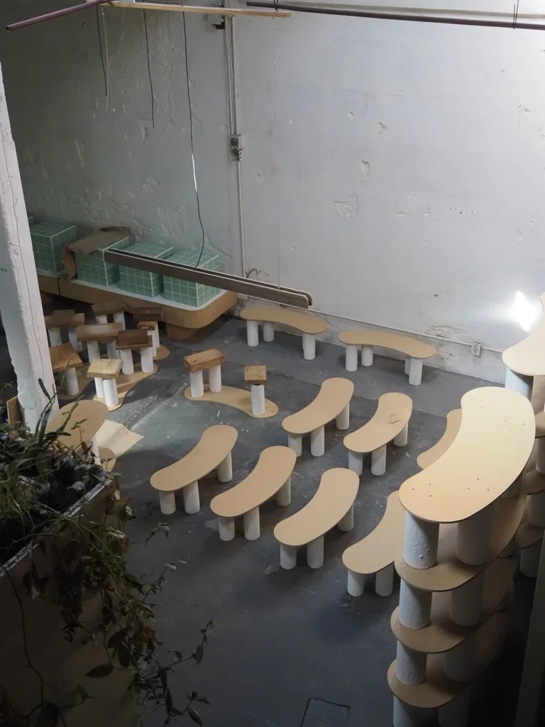 Multiple small wooden benches and tables arranged in a sequence, with a green tiled counter or shelf on the wall, in an industrial or workshop space with concrete flooring and unfinished walls.