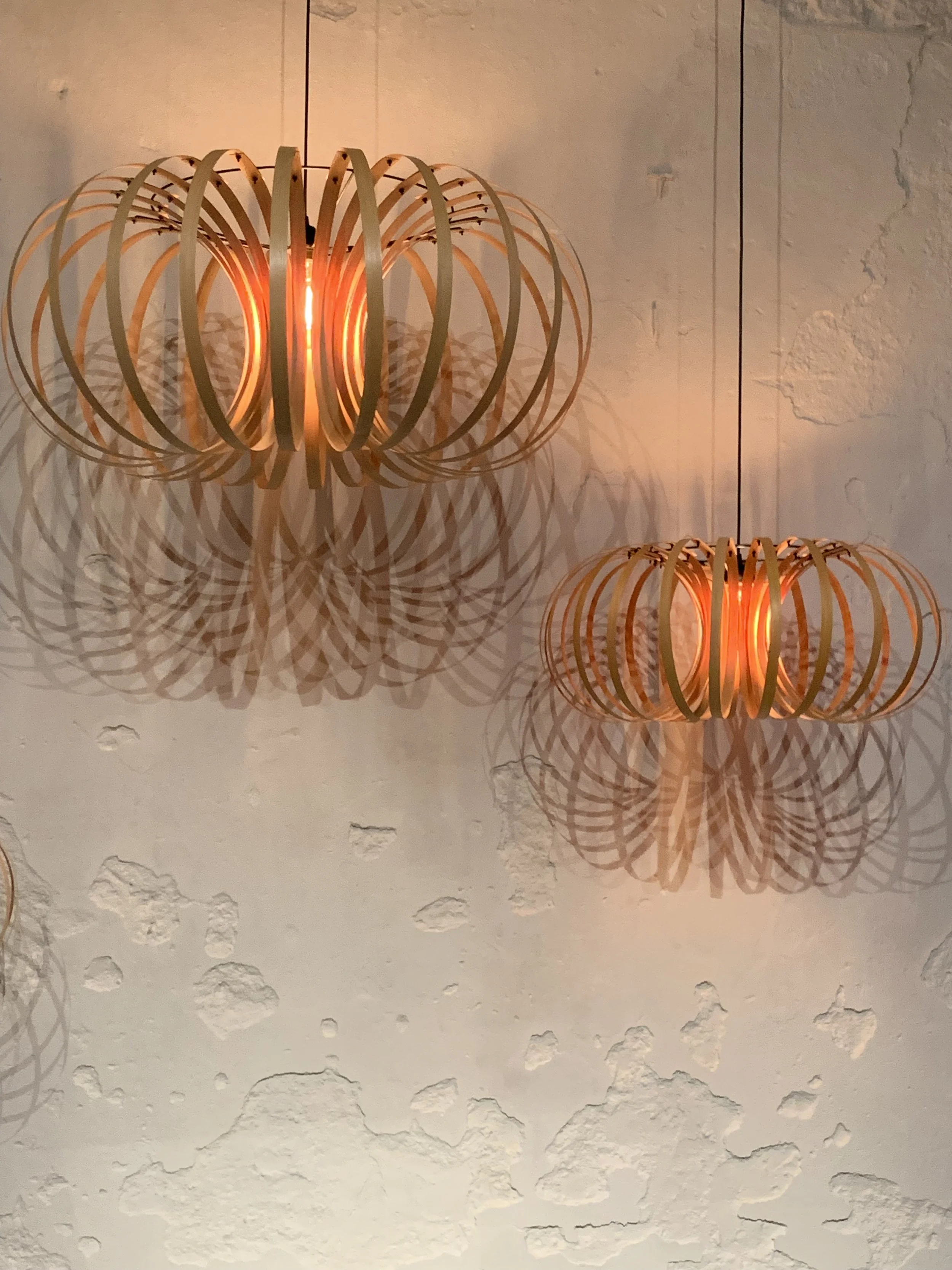 Two modern ceramic pendant light fixtures with wooden slats and glowing bulbs casting intricate shadows on a textured white wall.