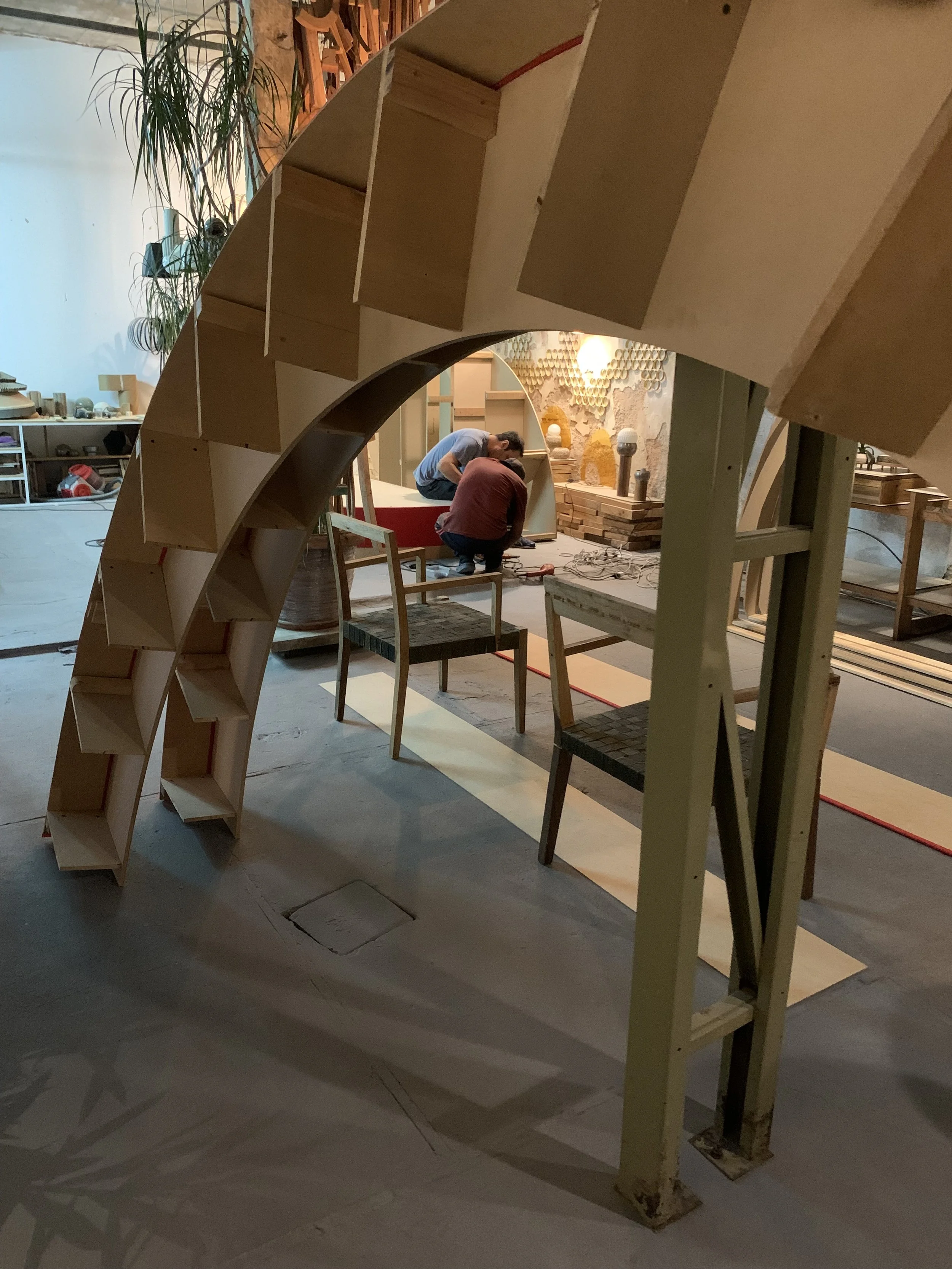 Two people working on a woodworking project inside a workshop, framed by a curved wooden structure with open shelving, surrounded by furniture and tools.