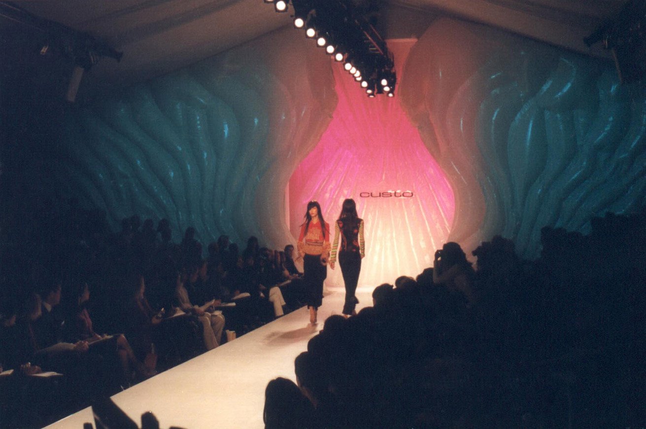 A fashion runway show with two models walking down the runway, showcasing colorful outfits. The background features a pink and blue artistic backdrop with the brand name 'Custo' displayed prominently.
