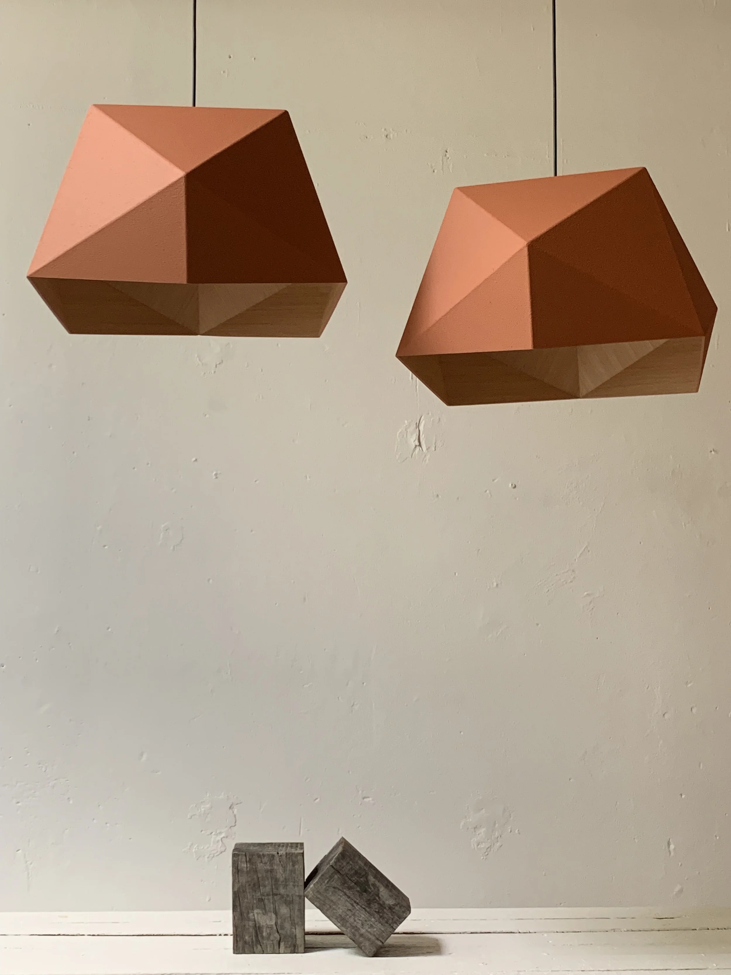 Two geometric pendant lights with pink and wooden surfaces hanging above two weathered wooden cubes on a white surface against a plain white wall.