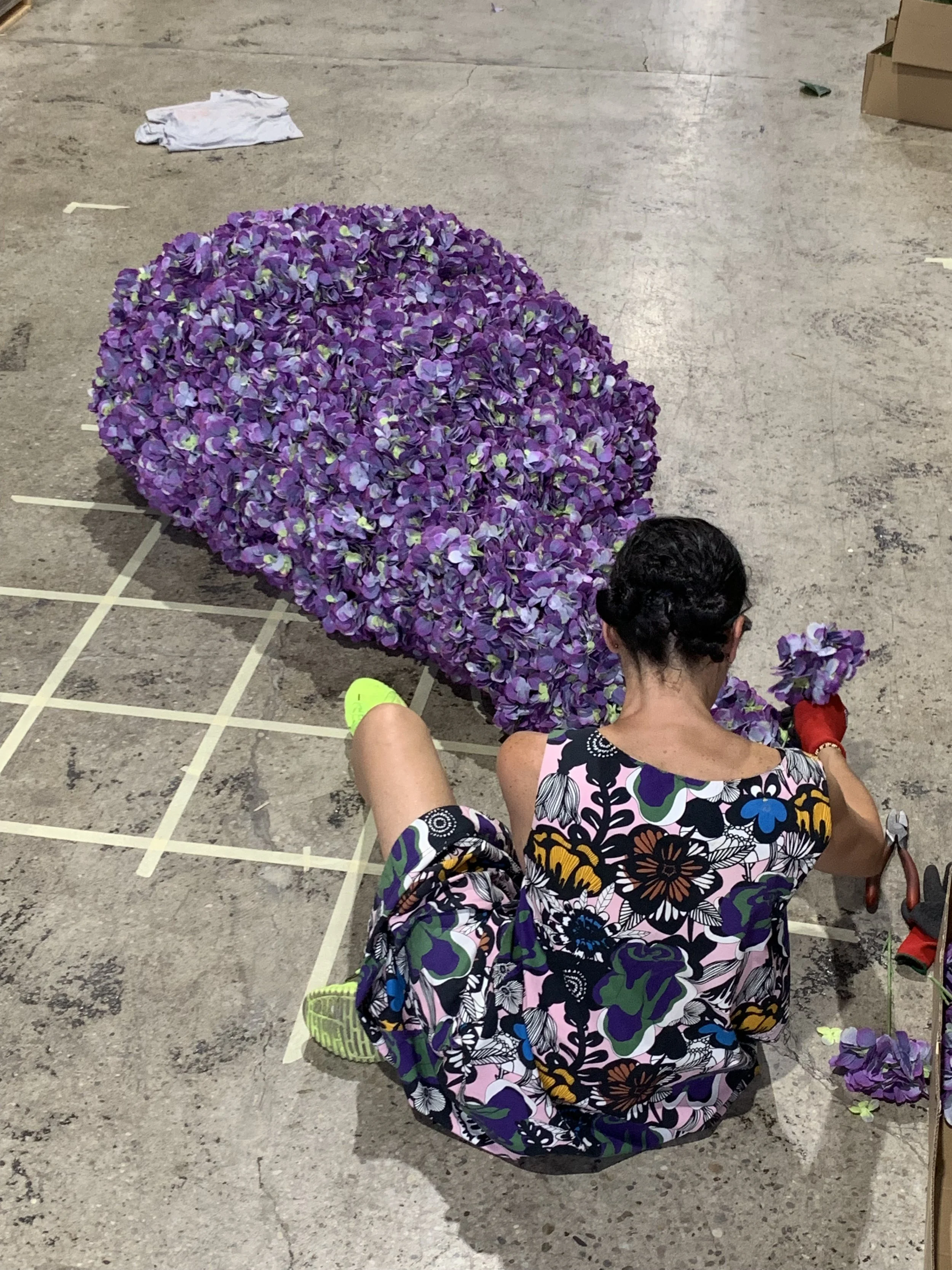 A woman seated on the ground arranging purple flowers, with a large bouquet or arrangement of purple flowers in front of her, on a concrete floor.