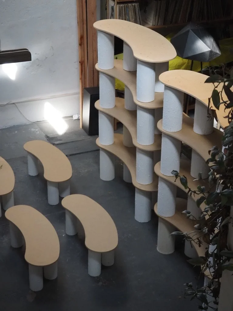 A multi-tiered modern wooden shelving unit with cylindrical legs, arranged in a curved pattern, located indoors near a white wall and a bookshelf.