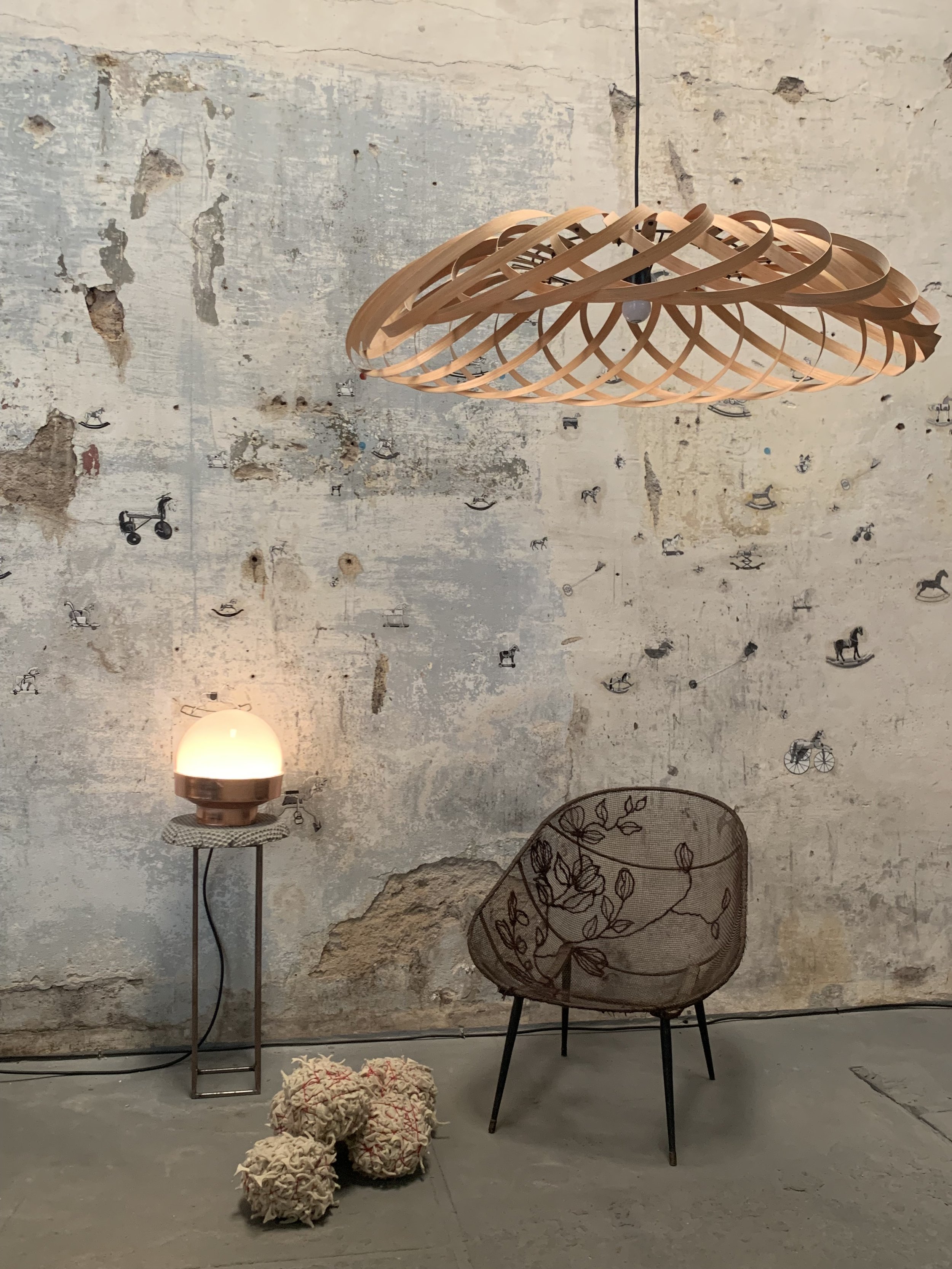 Interior with a rustic, weathered wall decorated with small horse and carriage illustrations, an abstract wooden pendant light, a vintage-style lamp on a stand, a wire chair with floral embroidery, and a rug with pom-pom embellishments on the floor.