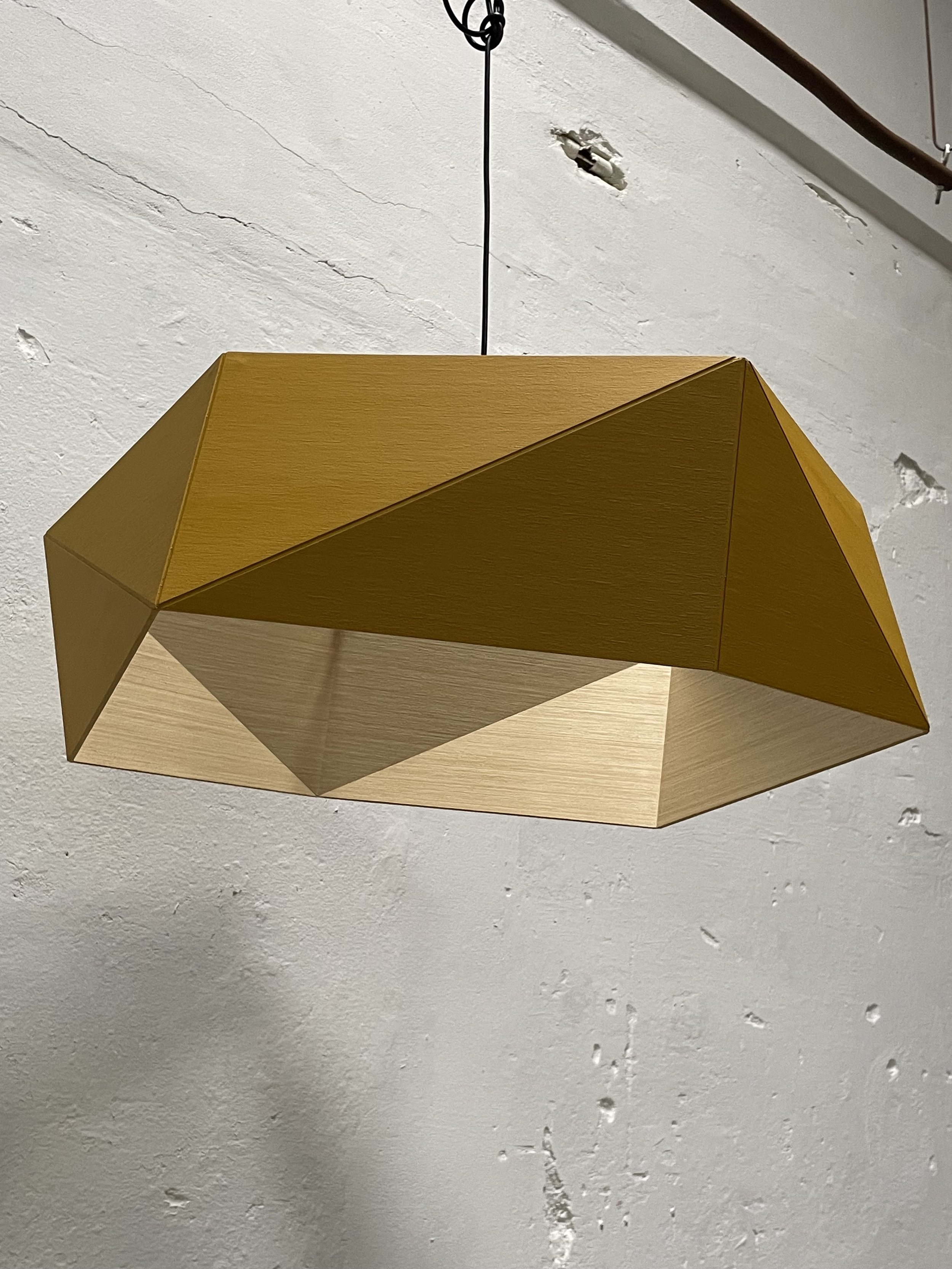 Modern geometric pendant light with wooden panels suspended from a black cord against a white textured wall.