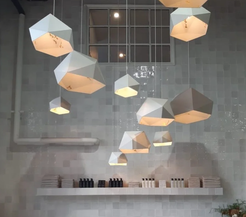 Multiple modern geometric pendant lights hanging from the ceiling above a shelf with stacked bottles and white towels against a tiled gray wall.