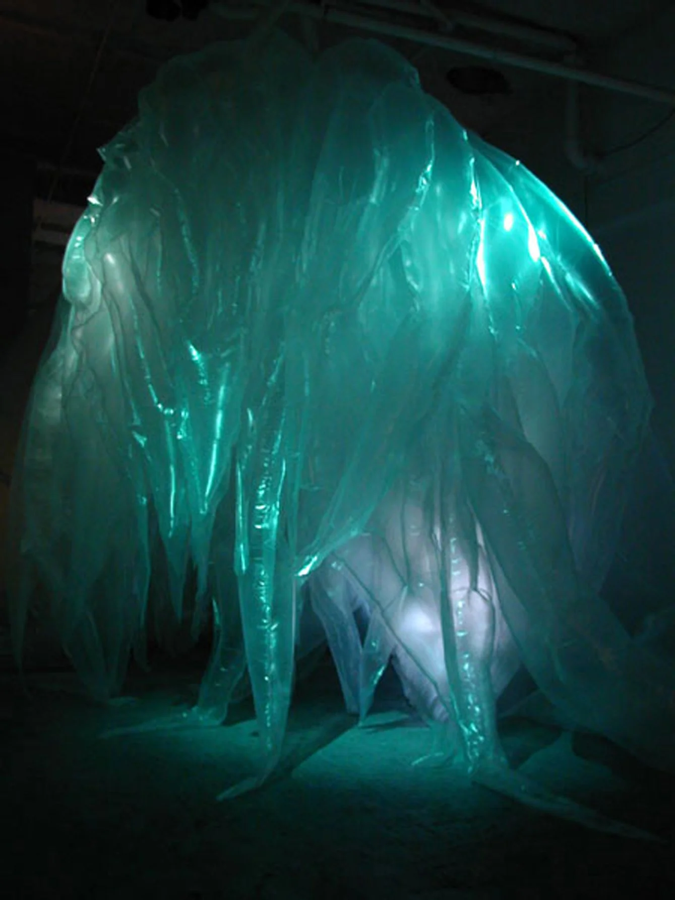Large illuminated sculpture made of translucent, glowing teal material resembling ice or a wave.