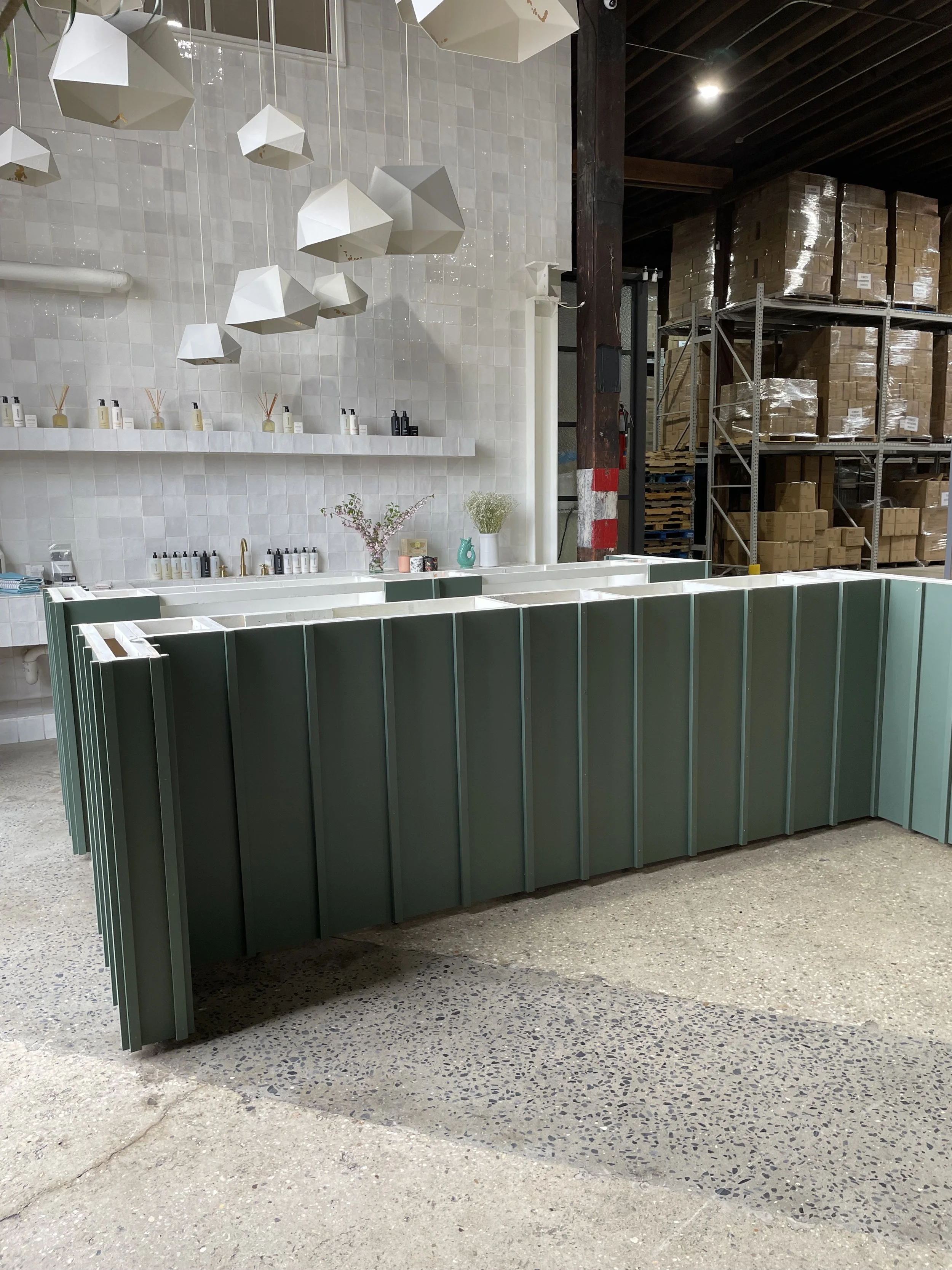 Interior of a store with a green display counter, white shelves with small containers, decorative vases, and dried flowers, white wall tiles, geometric pendant lights, and warehouse shelves with boxes in the background.