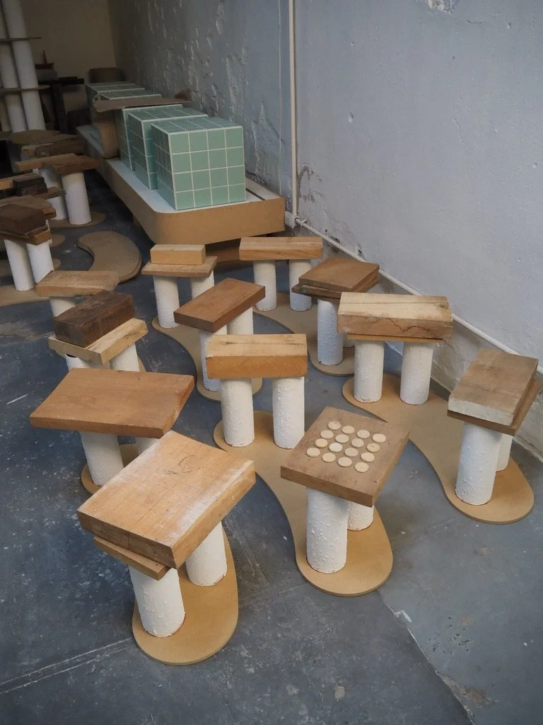 Multiple small tables with round white bases and square wooden tabletops, arranged in a semi-circle, with a green tiled display case or shelf in the background against a white wall.