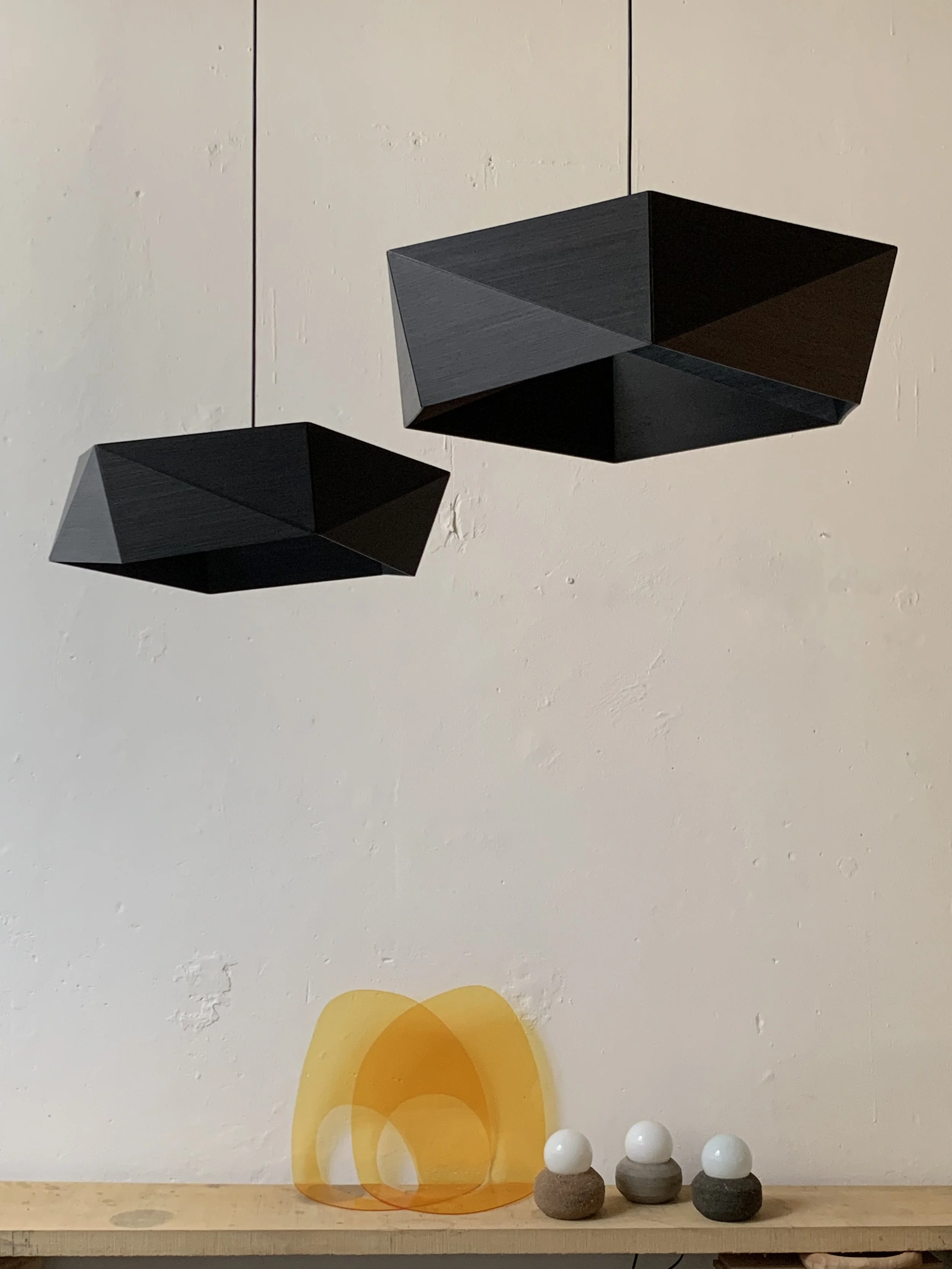 Decorative modern pendant lights with black geometric shapes hanging above a wooden surface. On the surface, there are three small decorative objects with white spheres on top of gray and black bases, and a yellow abstract art piece.