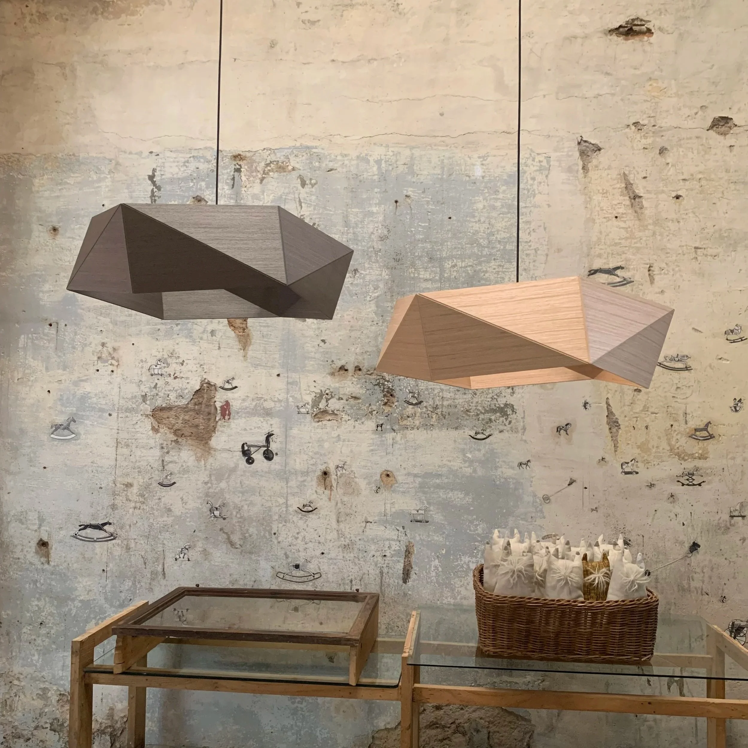 Two geometric pendant lights, one wood-colored and the other darker, hanging above a wooden table with a glass top and a basket filled with white fabric bags, against a distressed wall with small painted black and gray boats and animal figures.