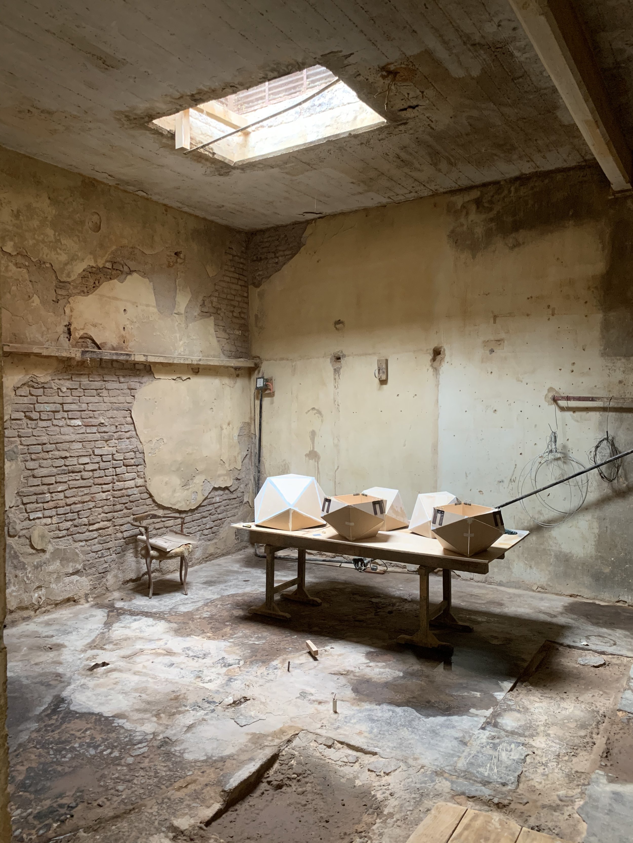 Interior of a room under construction with exposed brick and plaster walls, a large skylight, and a wooden work table with several geometric models on top.
