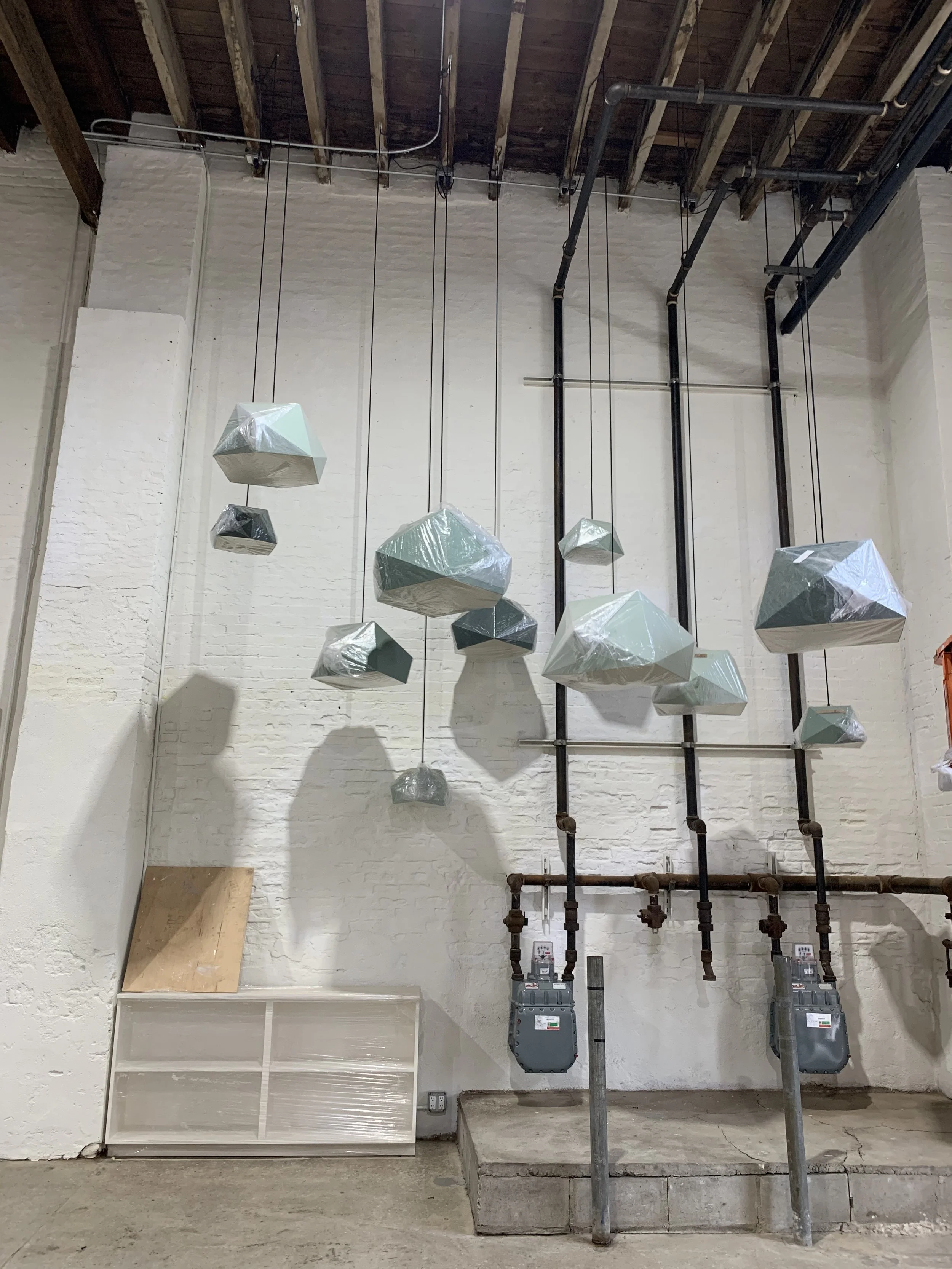 Industrial interior with hanging geometric-shaped light fixtures, pipes, and two electric meters mounted on the wall.