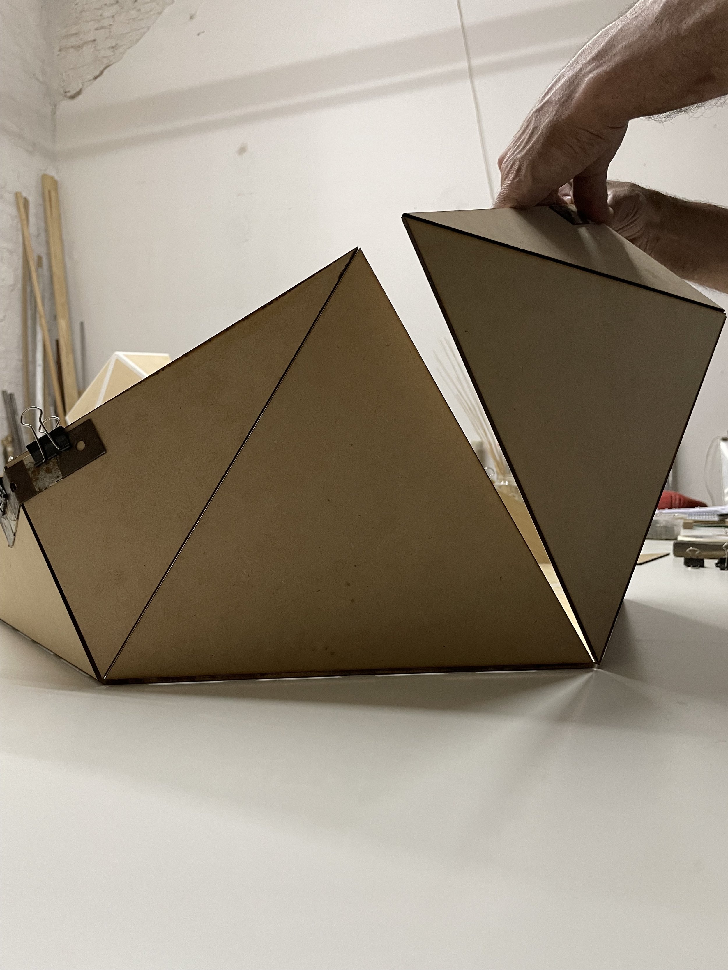 A person assembling a geometric wooden structure with triangular and rectangular shapes on a white surface in a workshop.