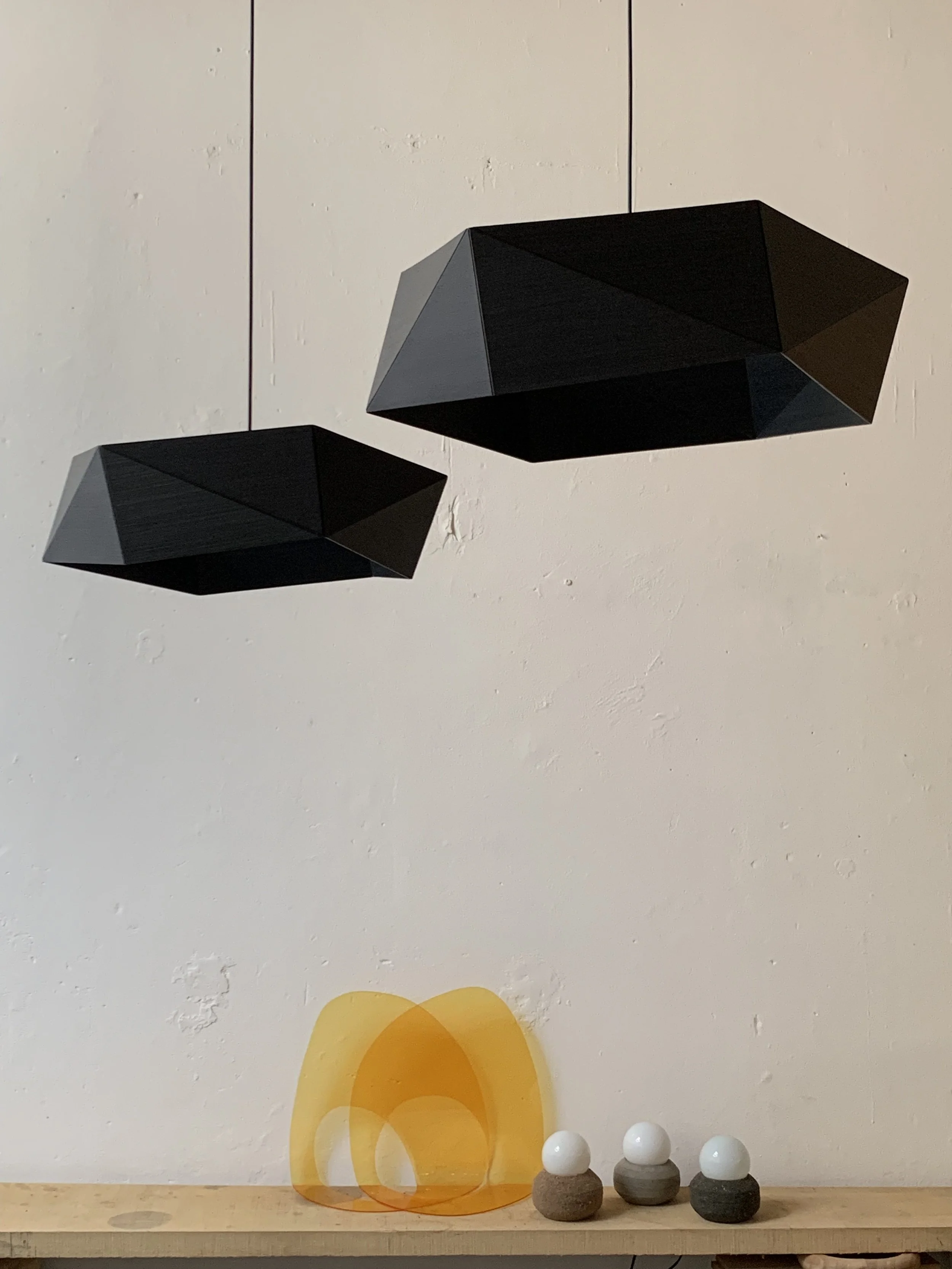 Two geometric black pendant lights hanging from the ceiling above a wooden countertop. On the countertop, there are two amber-colored and one yellow-colored abstract glass sculptures, along with three small rounded objects with white and gray tops and darker bases.