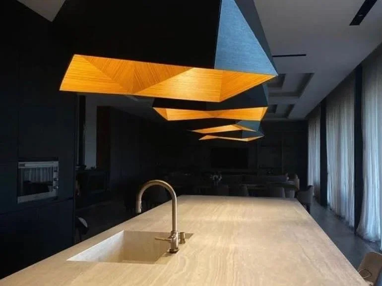 Modern kitchen with a beige countertop, built-in sink, and gold faucet, featuring a distinctive geometric wooden light fixture above, in a dimly lit room with large windows and curtains.