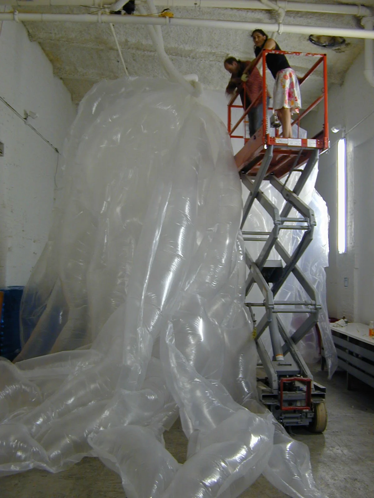 Two people on a scissor lift working on a large transparent plastic sculpture inside a warehouse or studio.
