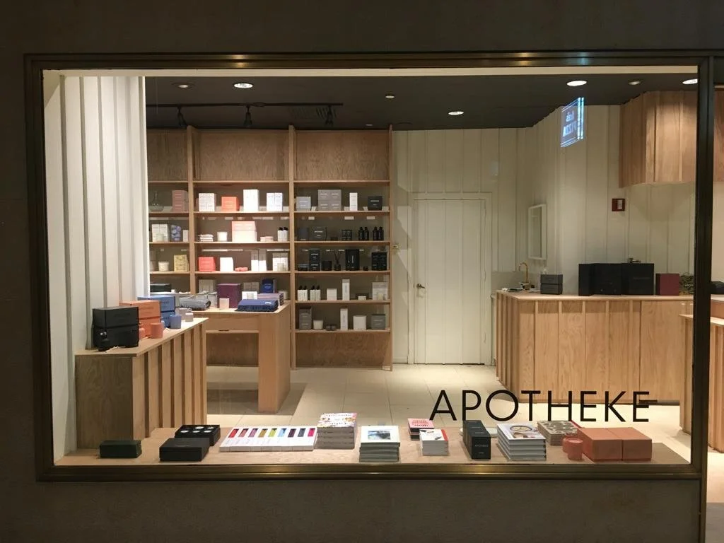 View of an apothecary shop through a large window, with wooden shelves and counters displaying various products such as boxes, bottles, and packages, and the word 'APOTHEKE' written on the glass.