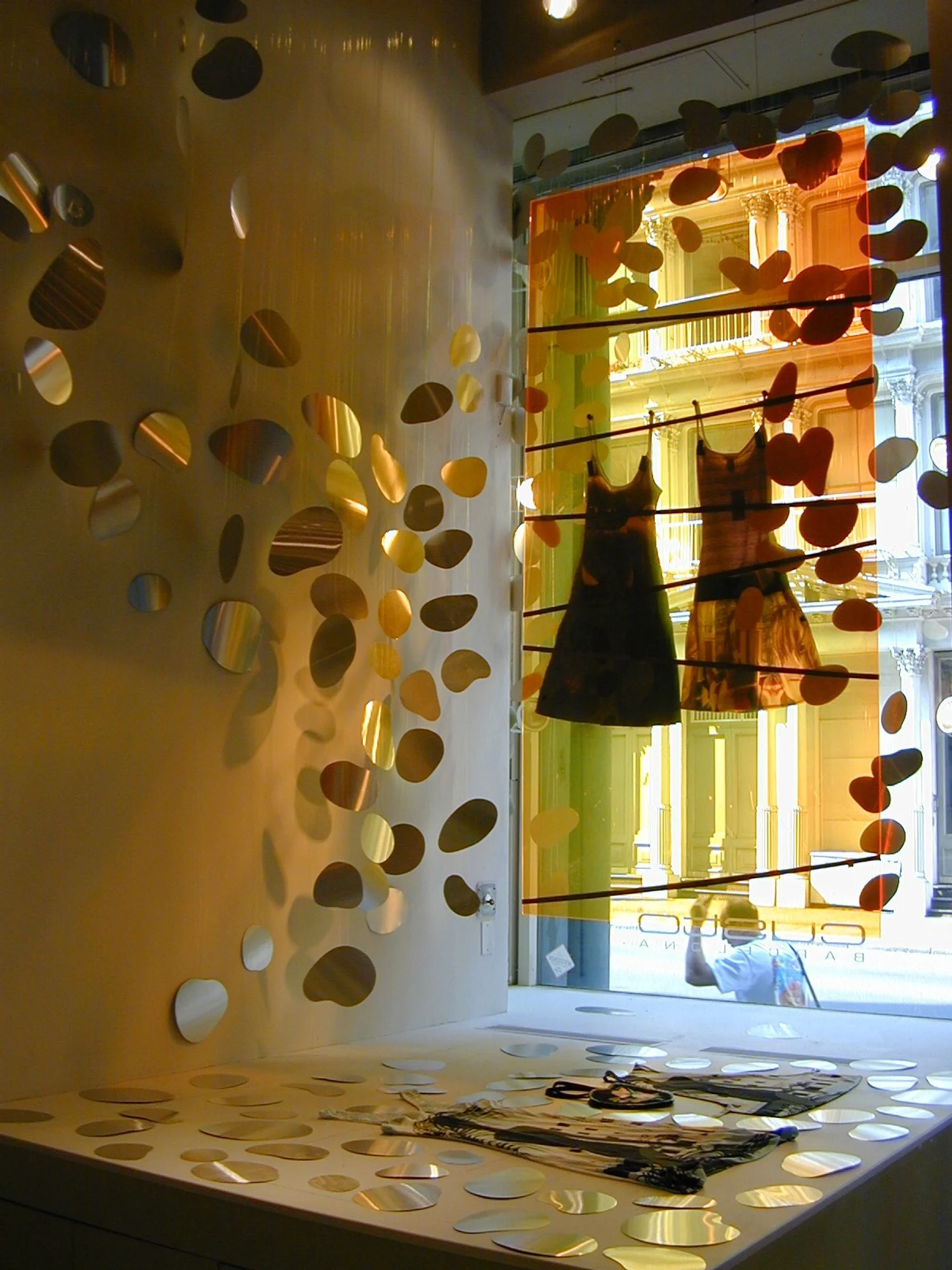 Display window with two dresses hanging, surrounded by circular reflective discs, with a view of a colorful building reflection outside.