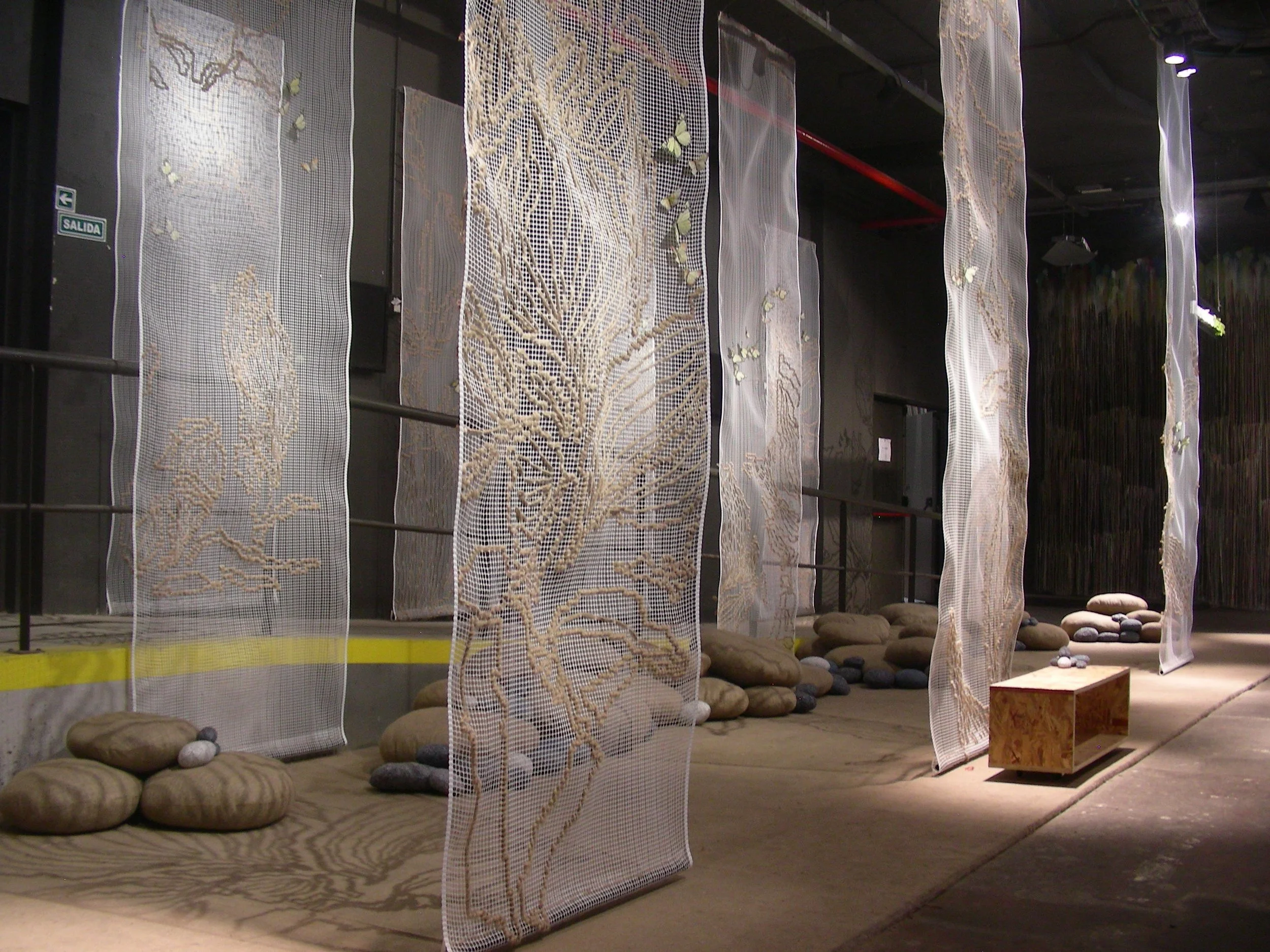 An indoor art installation features vertical fabric panels with embroidered tree and animal designs, surrounded by rocks and a wooden bench, in a dimly lit space.