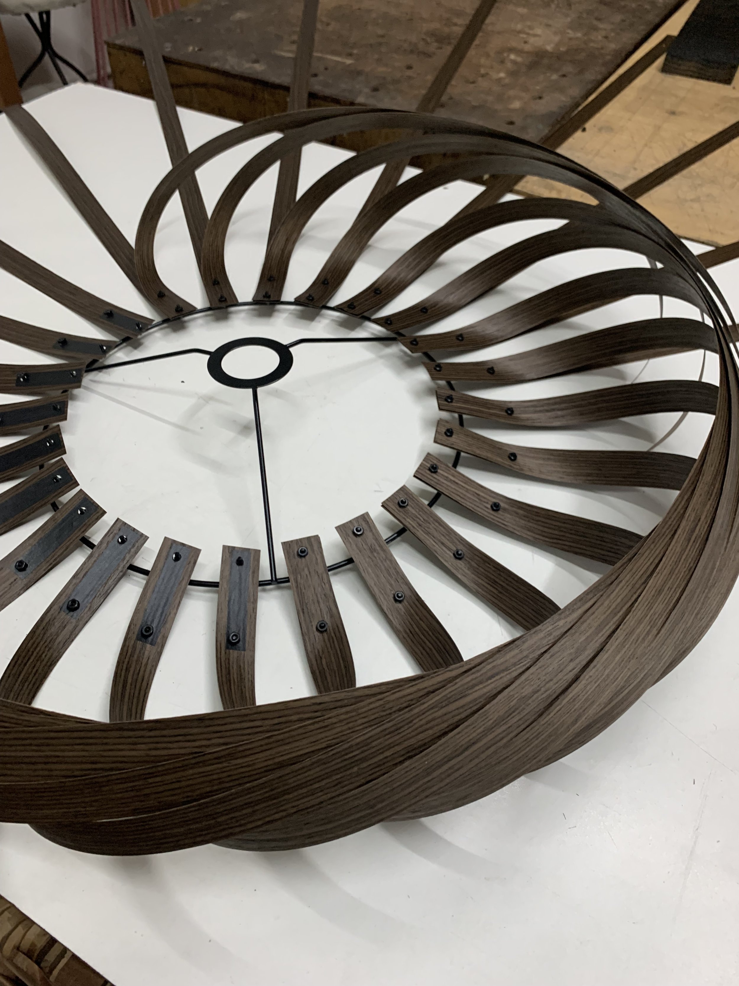 A modern wall clock with a spiral design made of wooden slats and black metal details, placed on a white surface.