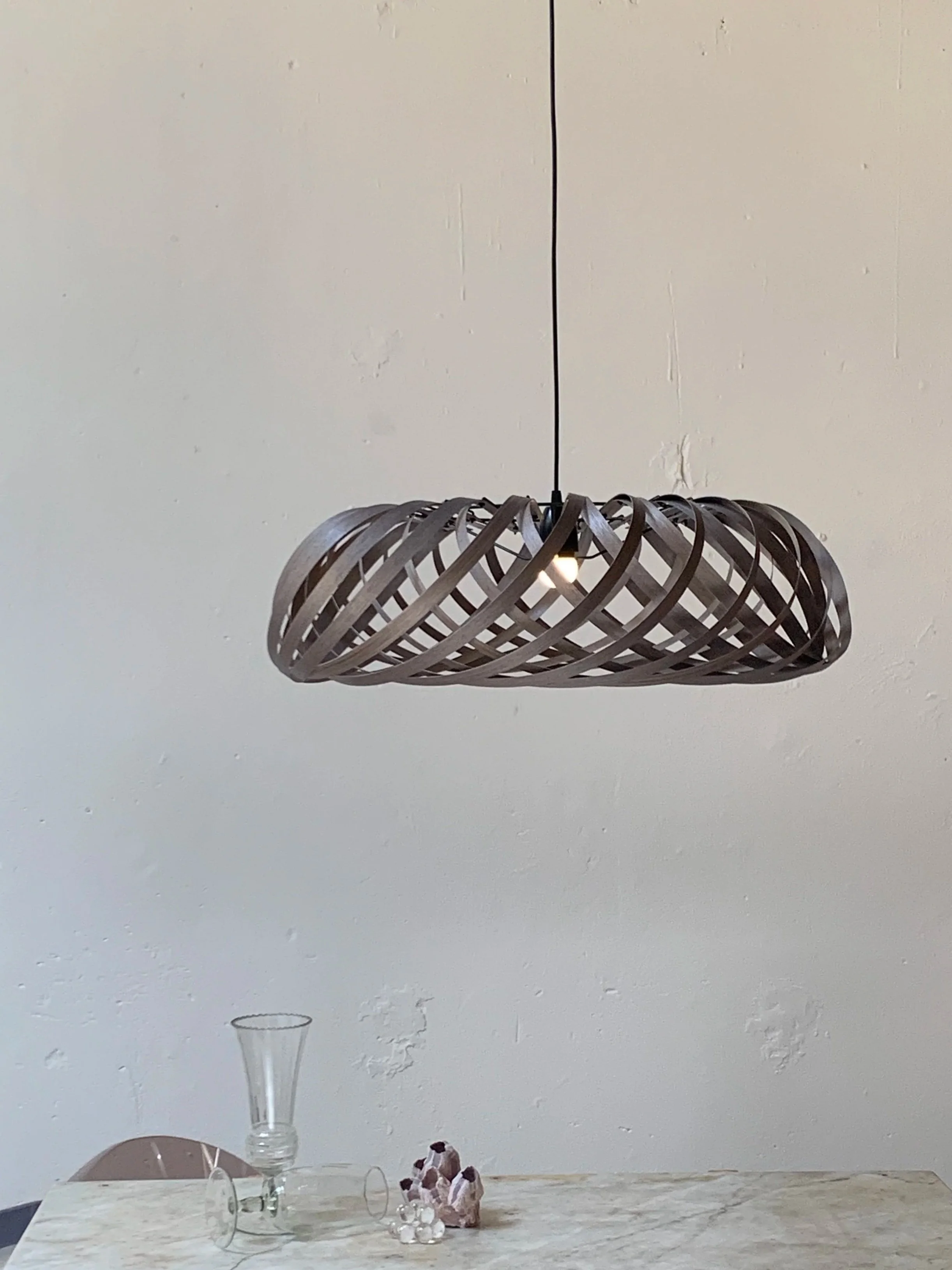 Modern pendant light with a swirling black metal design hanging above a marble table, with glassware and a small figurine on the table.