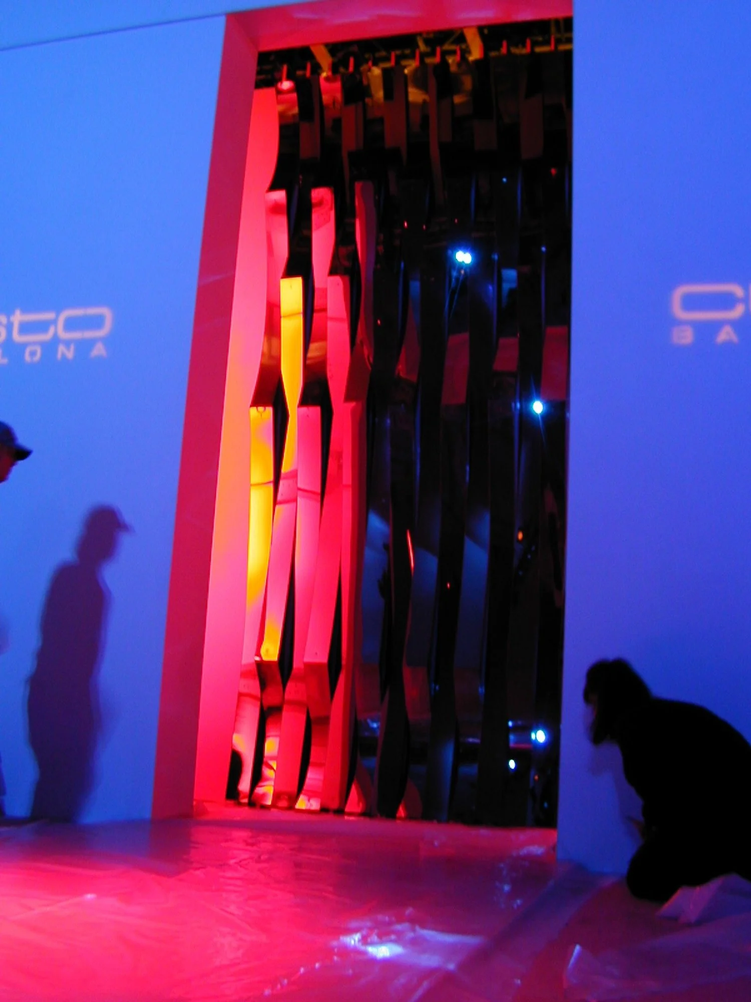 Interior of a store with colorful geometric architectural features illuminated with red, yellow, and blue lights, and shadows of people on the walls.