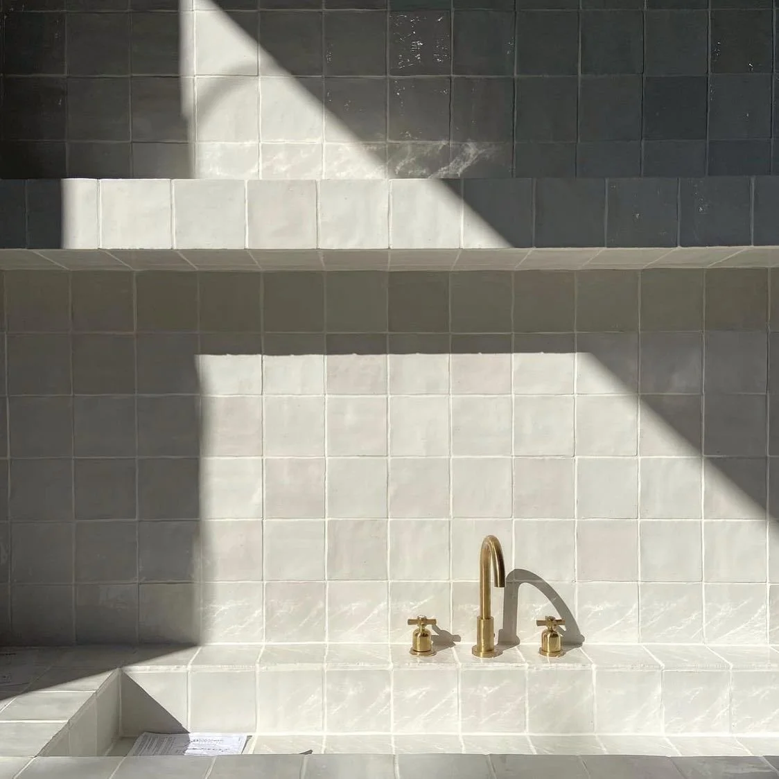 Close-up view of a tiled bathroom or shower wall with a gold faucet and handles, with sunlight casting shadows on the tiles.