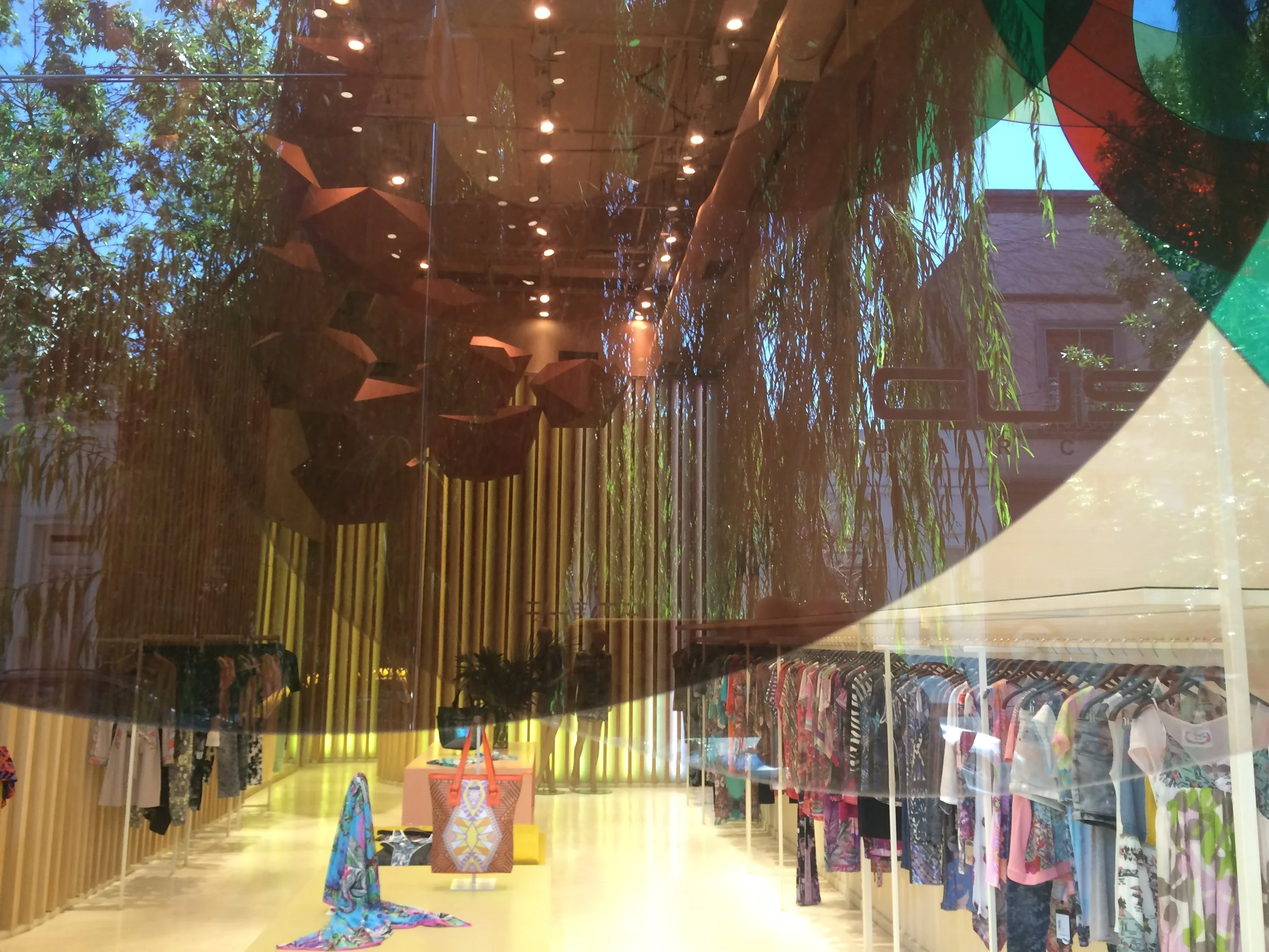 Clothing store window display with colorful garments on racks, decorative umbrellas hanging from the ceiling, and reflections of trees and the street outside visible through the glass.