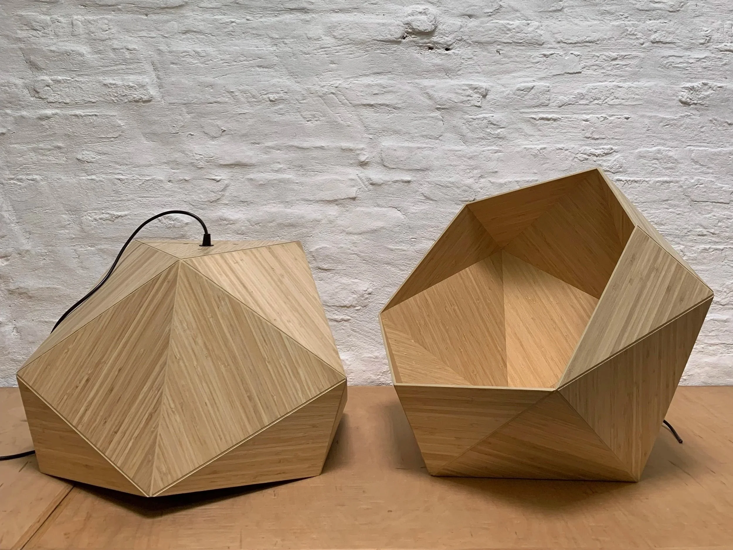 Two geometric wooden lamps with polygonal shapes and a natural wood finish, placed on a wooden surface against a white brick wall background.