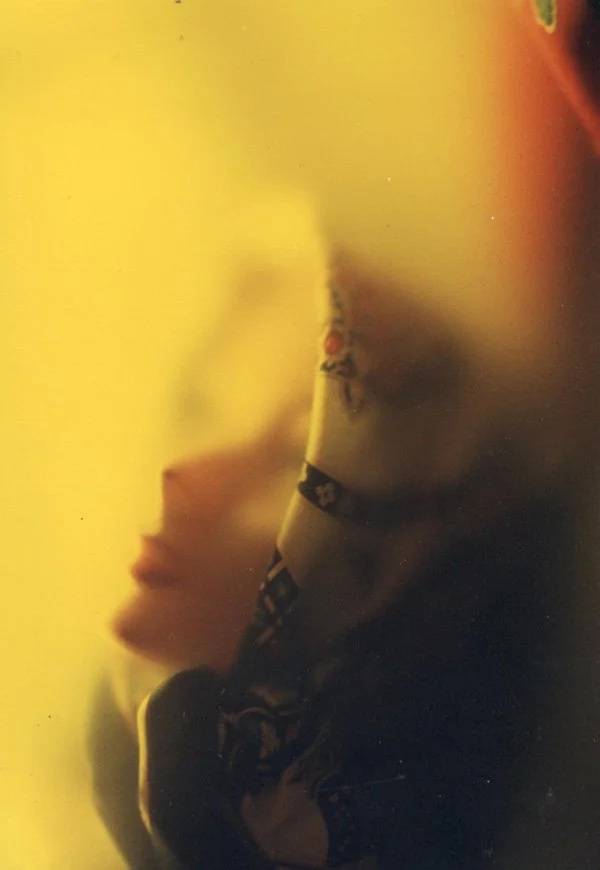 A side profile of a woman with dark curly hair, wearing a dark top, in front of a yellow background, looking away from the camera.