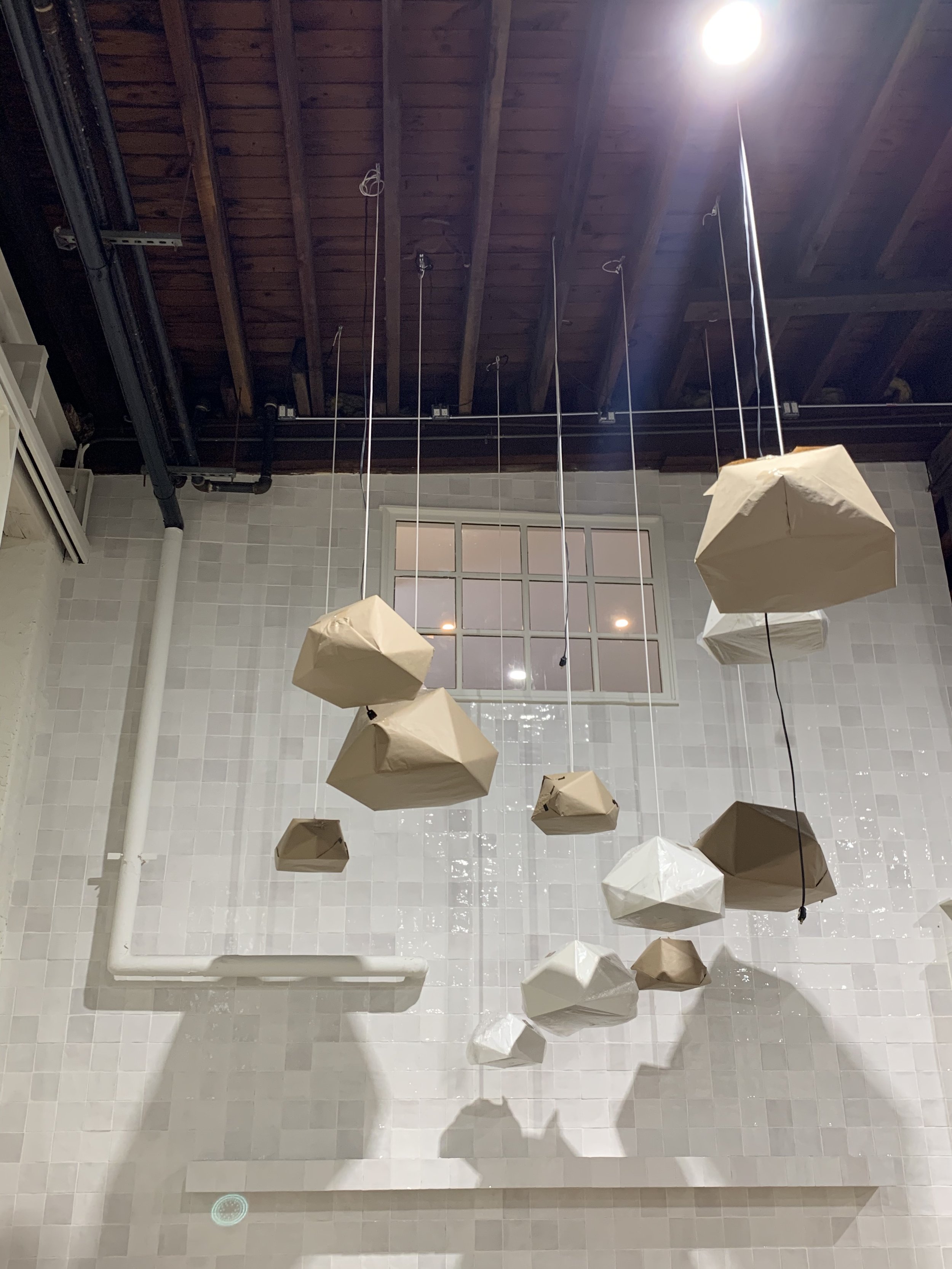 Hanging paper lanterns in various shades of beige and white, suspended from a wooden ceiling with visible beams, in front of a tiled white wall with a window.