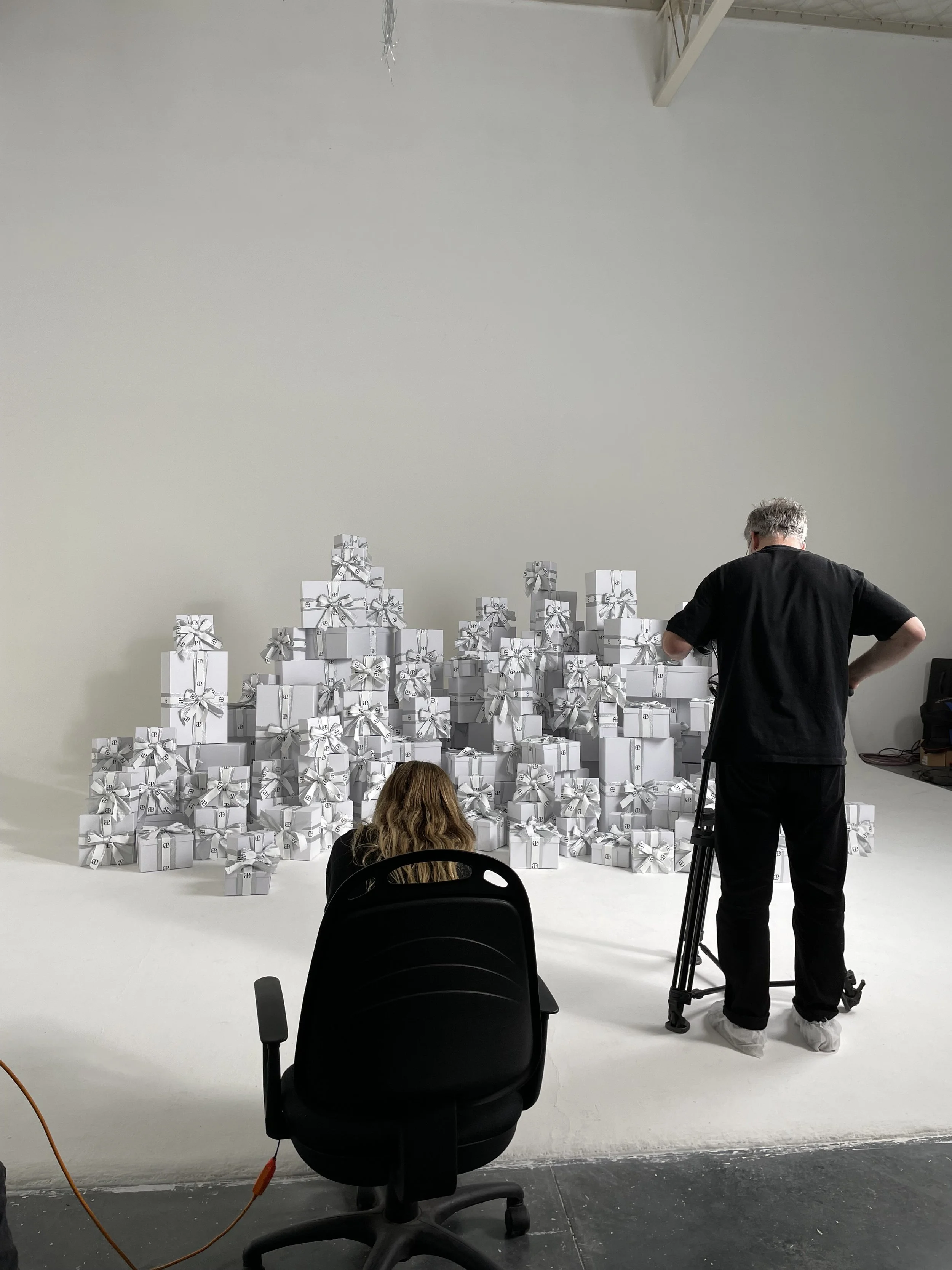 Studio scene with a man and woman in front of a wall of white gift boxes with silver ribbons, woman sitting in a chair and man operating a camera.