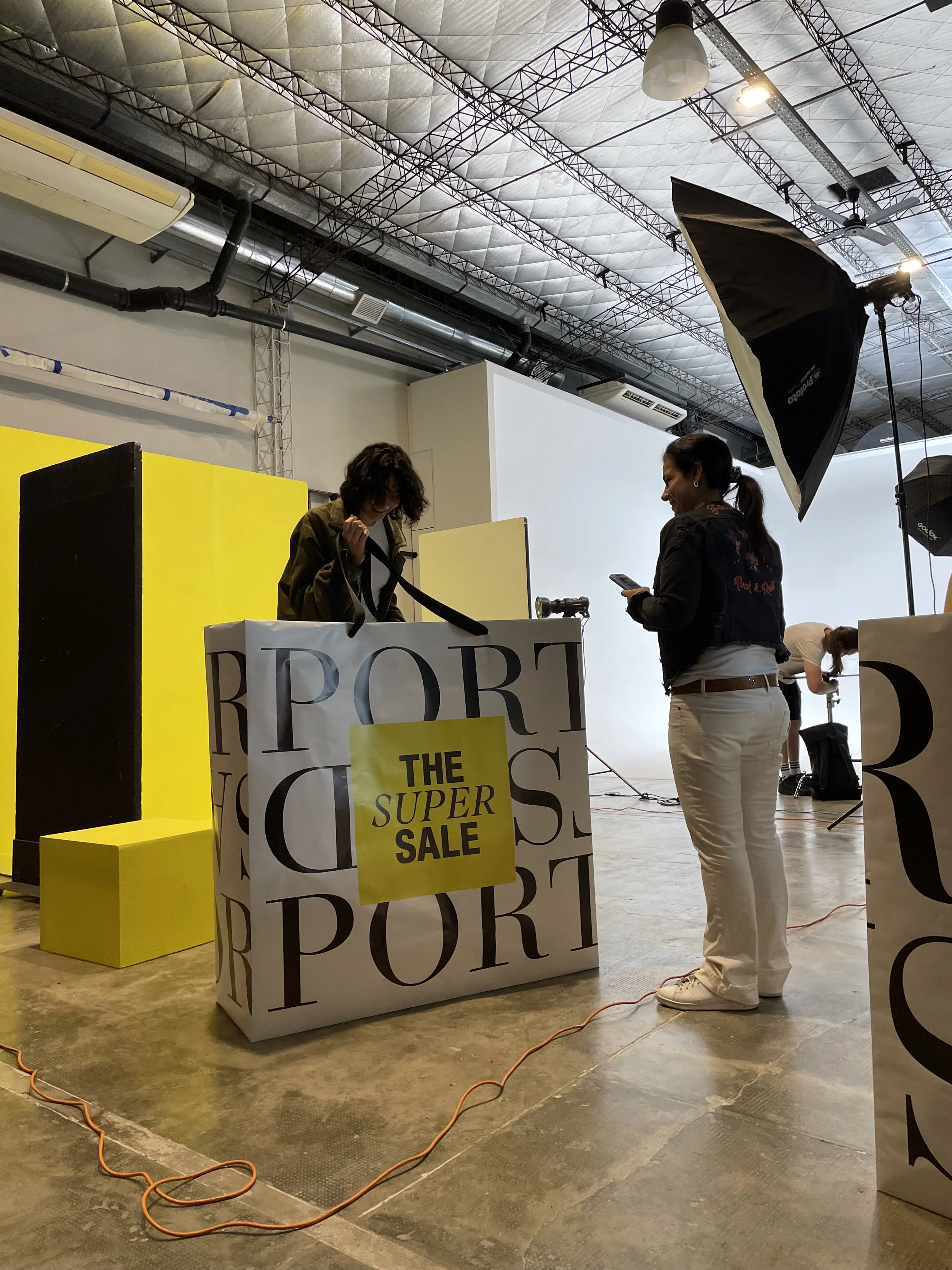 Two women are in a photography studio with a large white backdrop and studio lights, preparing for a shoot with a festive sale sign reading 'The Super Sale' in yellow and black.