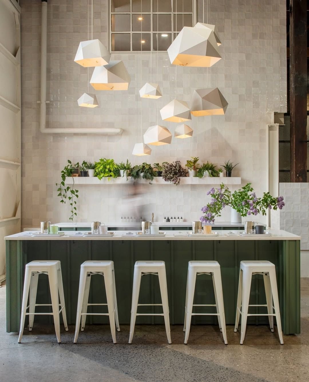 Modern kitchen with a white countertop island, green base, four white barstools, hanging geometric white pendant lights, and a wall shelf with various green plants.
