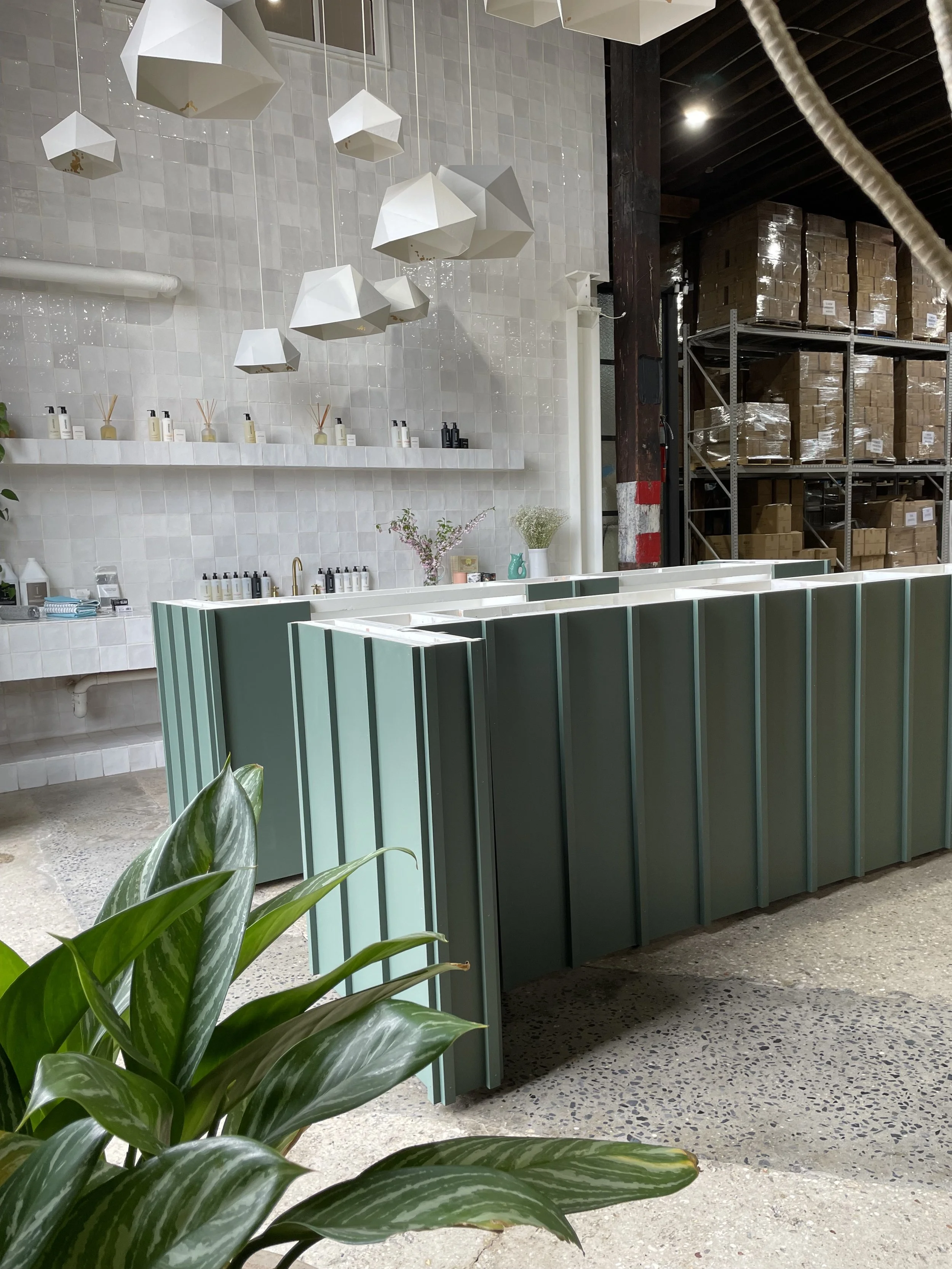 Interior of a modern retail or showroom space with hanging geometric pendant lights, white tiled wall with shelves of bottles, a green panel counter, potted plants, and industrial shelving with boxes in the background.
