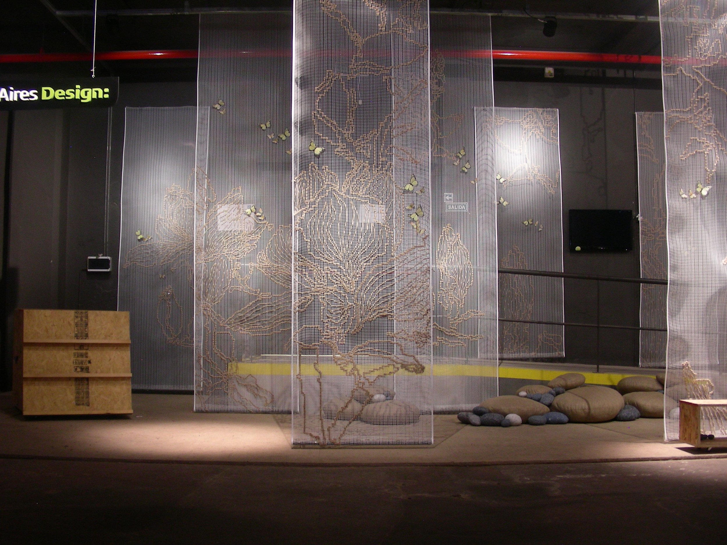 Interior view of a modern art installation featuring large fabric panels with floral patterns suspended from the ceiling, decorative rocks on the floor, a black television on the wall, and a small wooden crate on the ground.