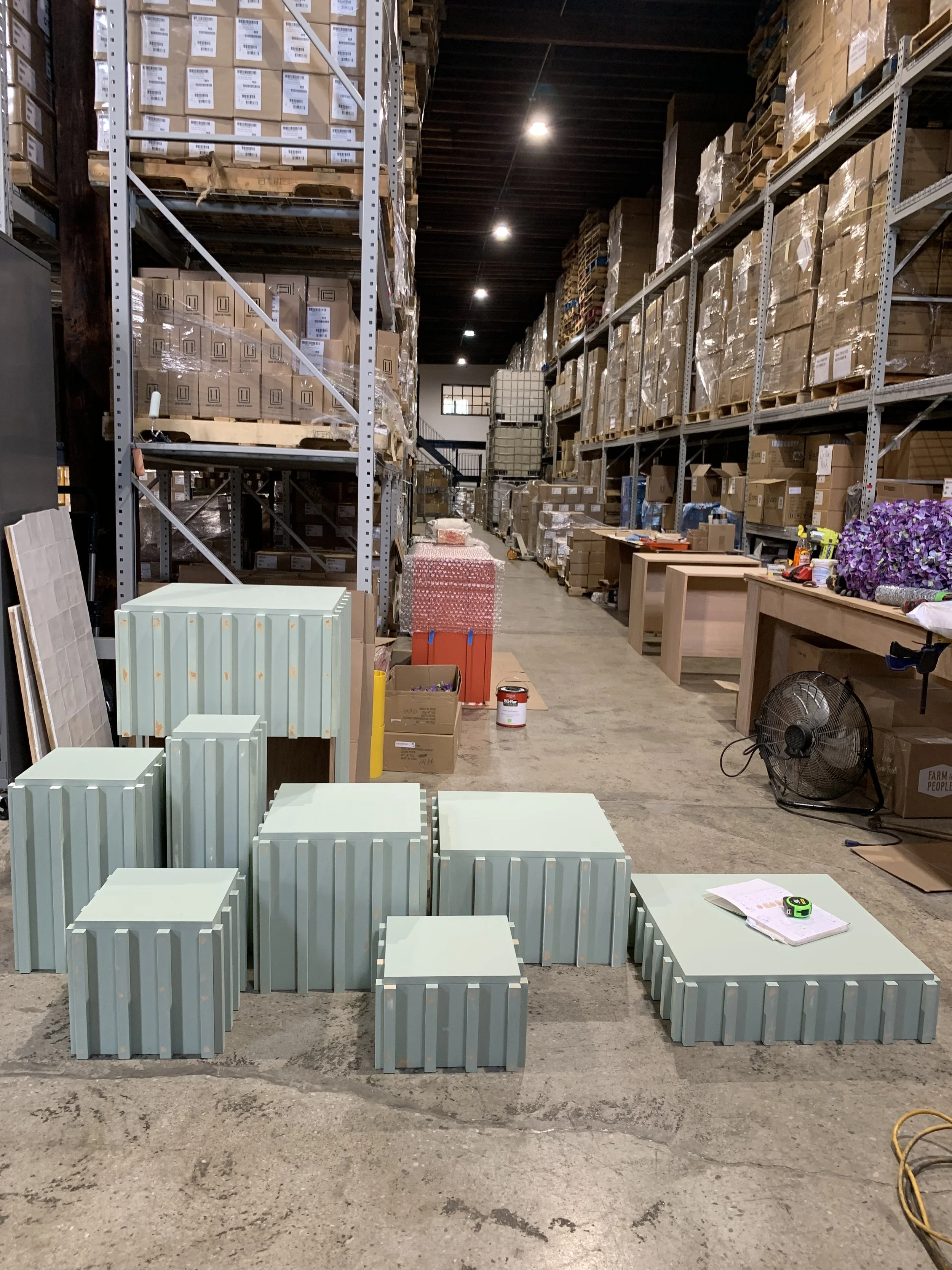 Warehouse with metal shelves filled with boxes, some on pallets. On the floor, there are multiple light green metal boxes or containers of varying sizes, a notebook, a tape measure, and some papers. In the background, a table holds tools and a fan is