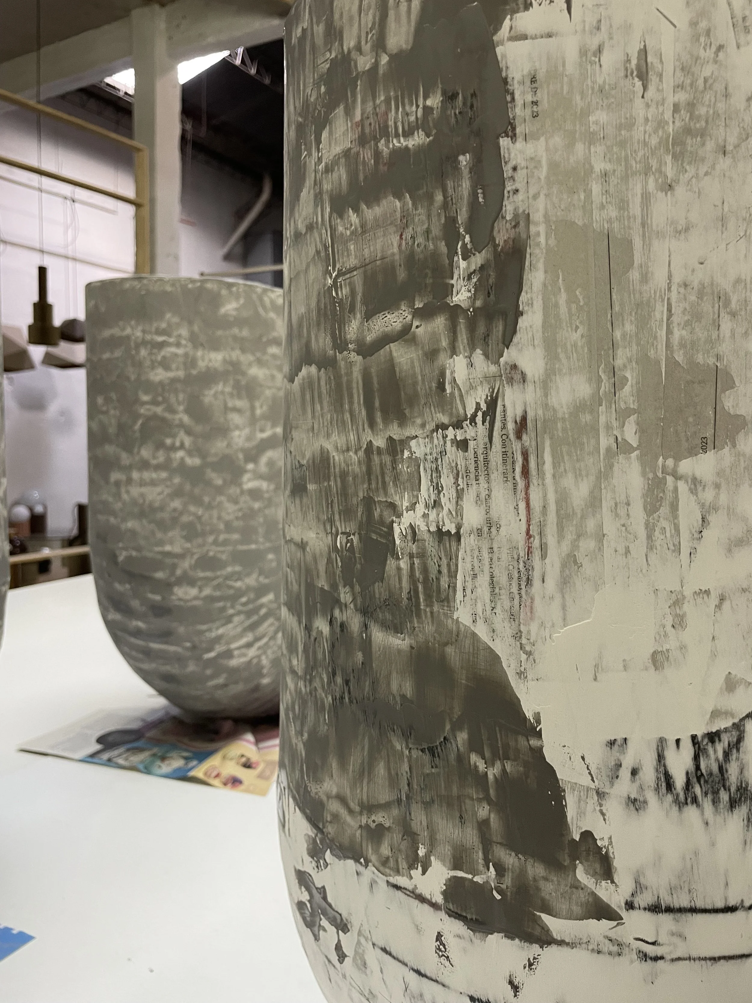 Close-up of an unfinished ceramic or plaster vessel with a rough, painted surface, in a workshop setting. In the background, there is another similar vessel, some magazines or catalogs, and workshop tools or shelves.