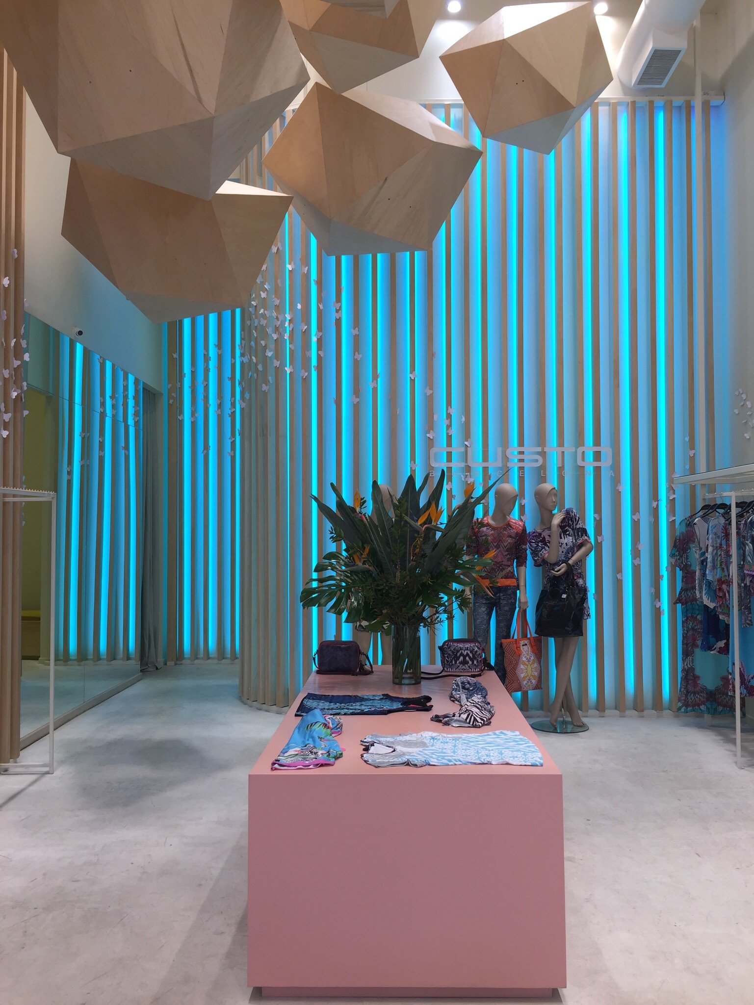 A retail store display with a pink table showcasing clothing and accessories, mannequins dressed in colorful outfits, a large plant centerpiece, and a background of vertical blue lighting, wooden geometric ceiling fixtures, and white butterfly decora