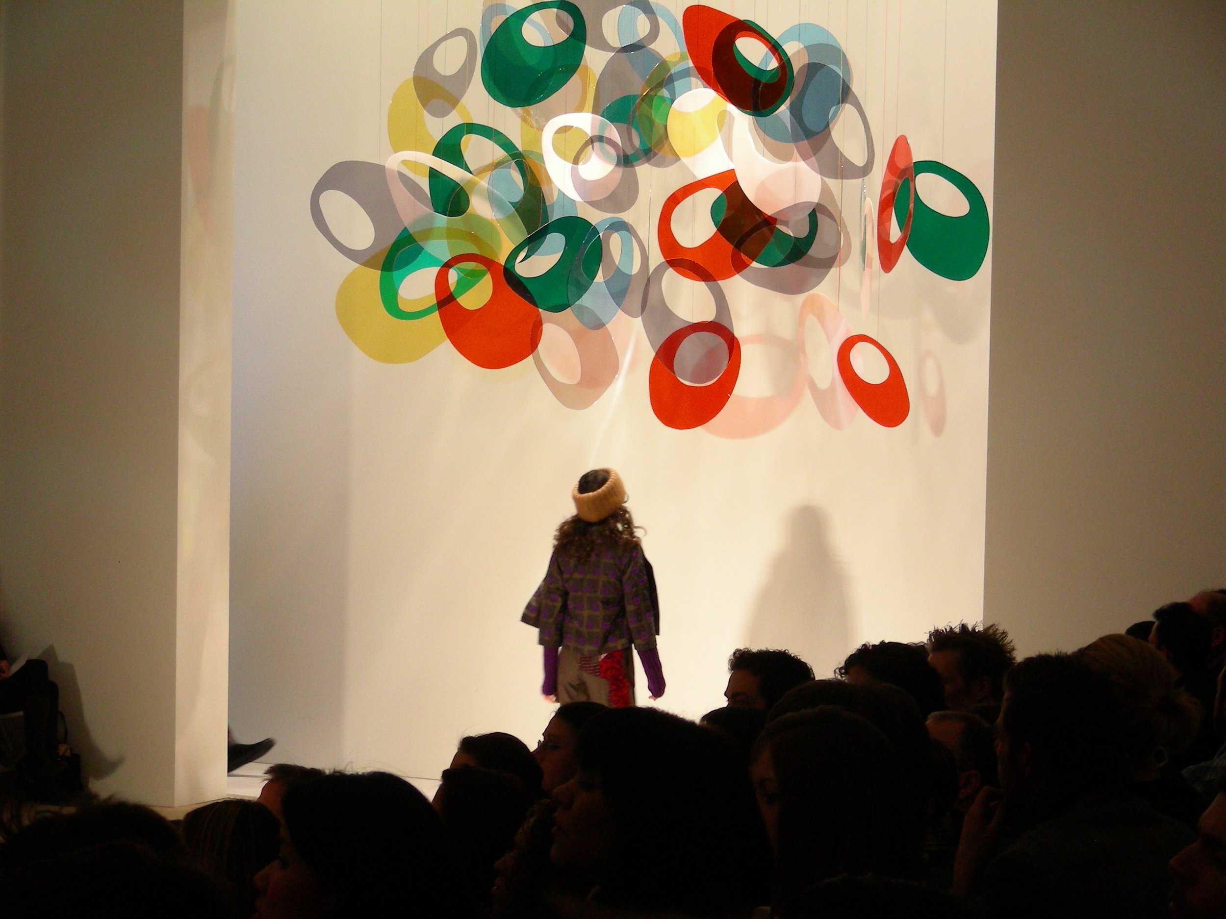 A woman in a purple plaid jacket and beige hat looking at a colorful abstract sculpture of overlapping red, green, yellow, and gray oval shapes on a white gallery wall, with an audience watching.