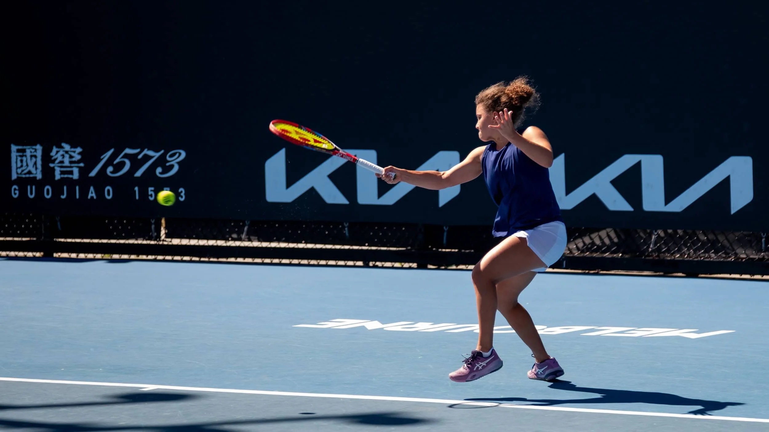A young girl with curly hair playing tennis on an outdoor court, mid-action hitting a tennis ball with a racket, wearing a blue sports top, white shorts, and sneakers, with a dark background featuring large text and Chinese characters.